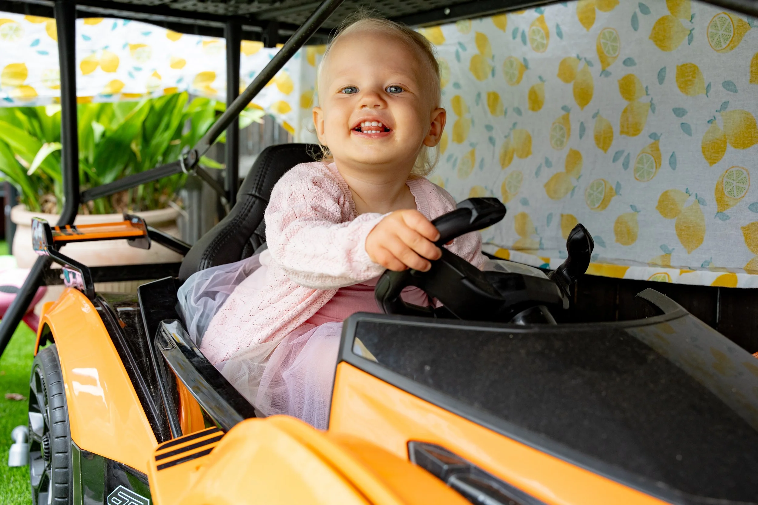 A young girl with blond hair wearing a pink dress and a light pink cardigan sitting in a yellow toy car, smiling and holding the steering wheel, outdoors with green plants and a lemon-patterned background.