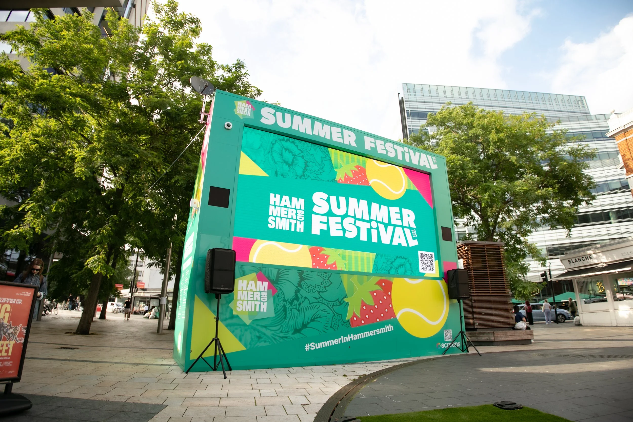 A large outdoor digital screen displays an advertisement for the Hammersmith Summer Festival in 2023. The screen is set up in a city street with trees and modern buildings in the background.