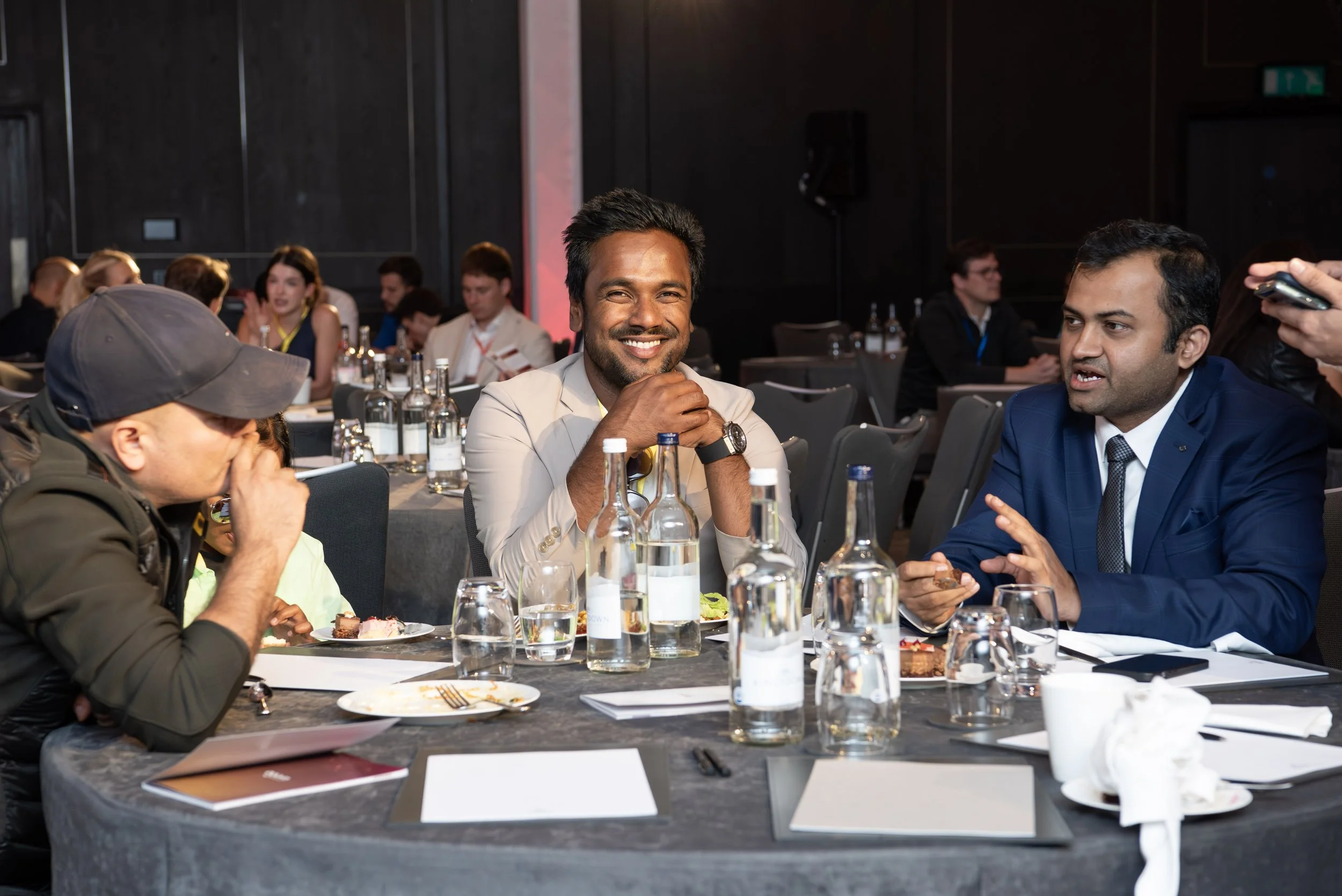 People sitting at a round table at a conference or event, engaging in conversation, with refreshments and documents on the table.
