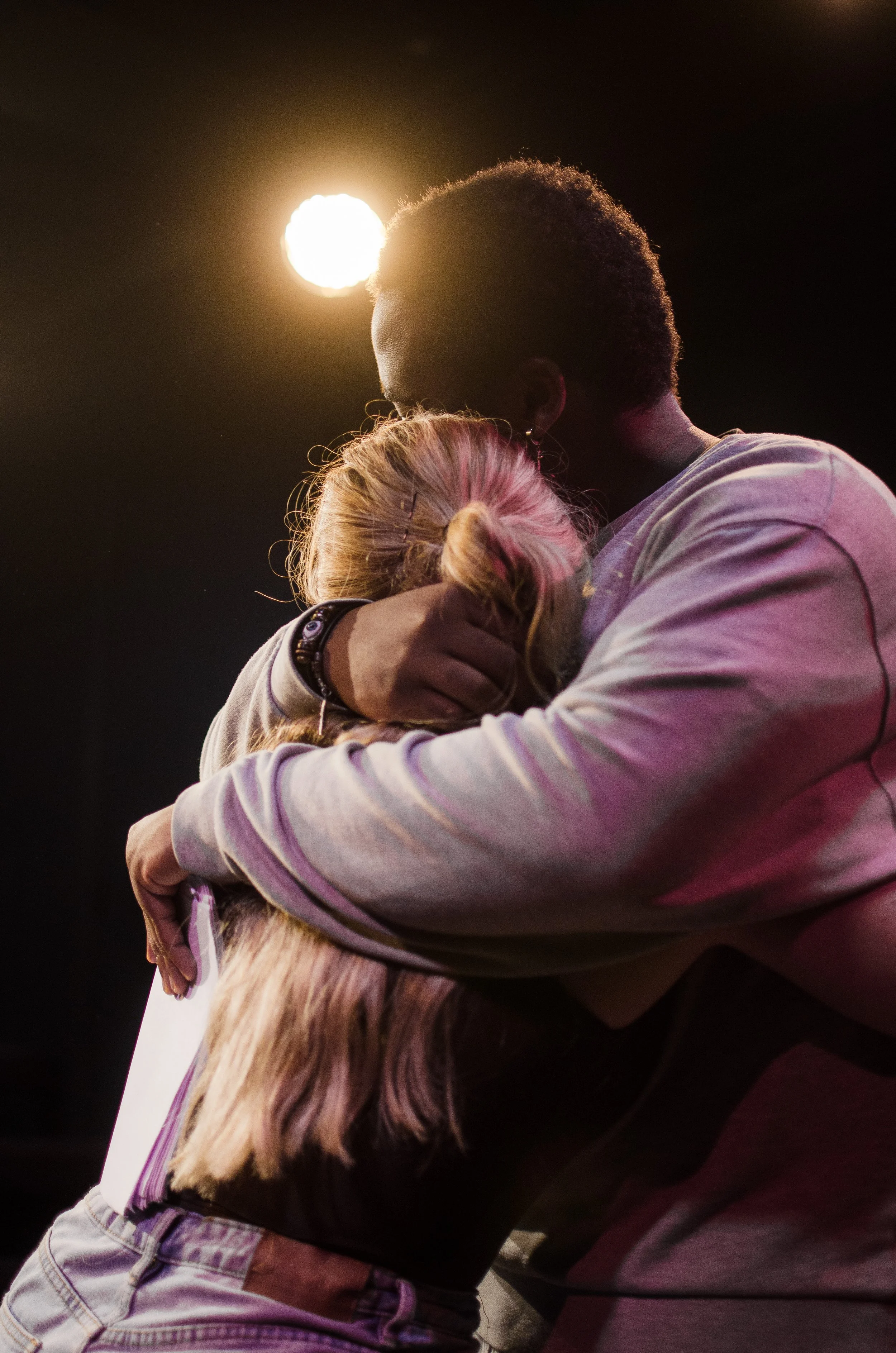 A man and a woman embrace in a hug, with the man’s head resting on the woman’s head under a bright overhead light.