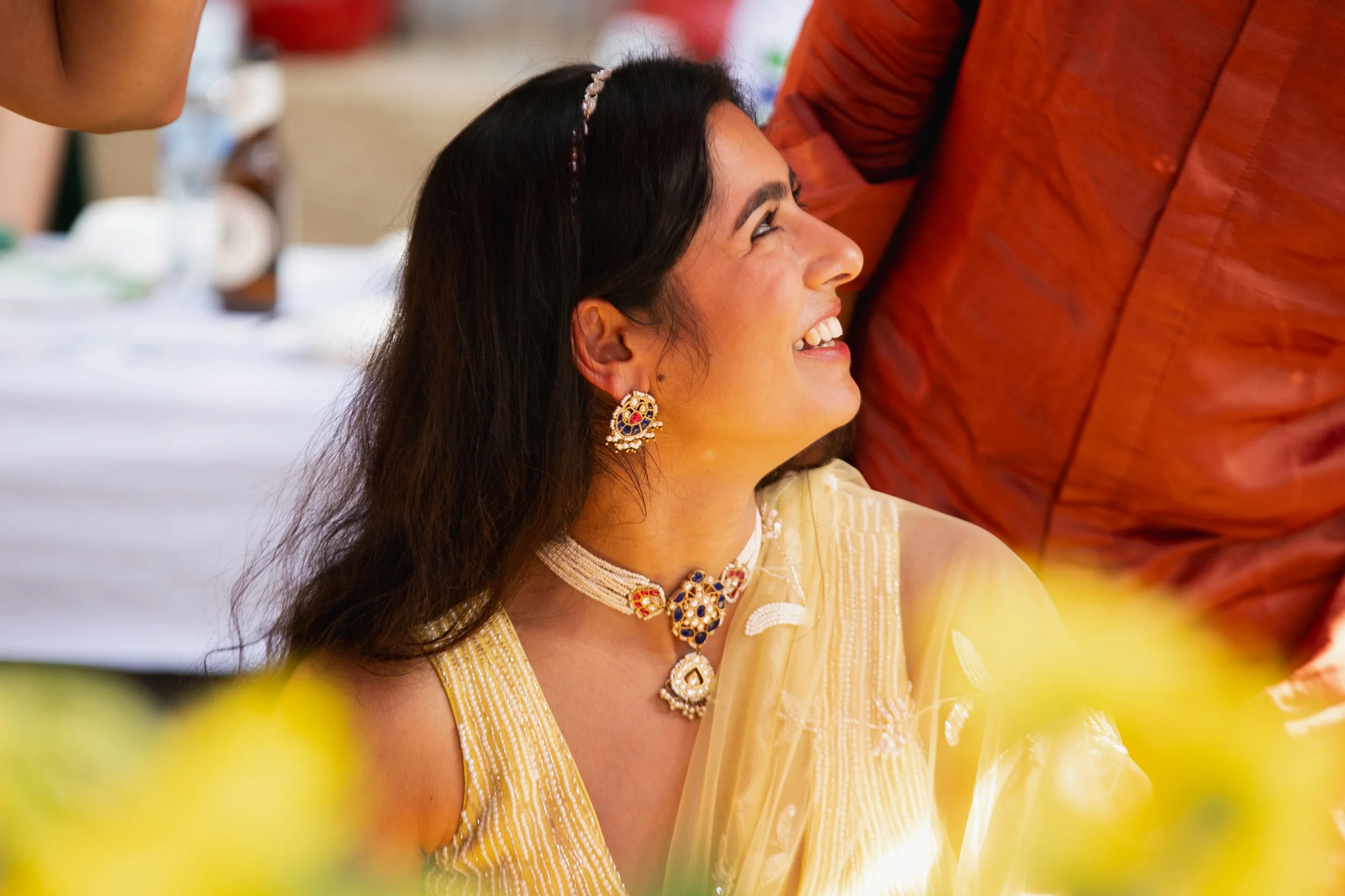 A woman dressed in yellow traditional Indian attire with jewelry, smiling and looking at a person near her at a celebration or wedding event.