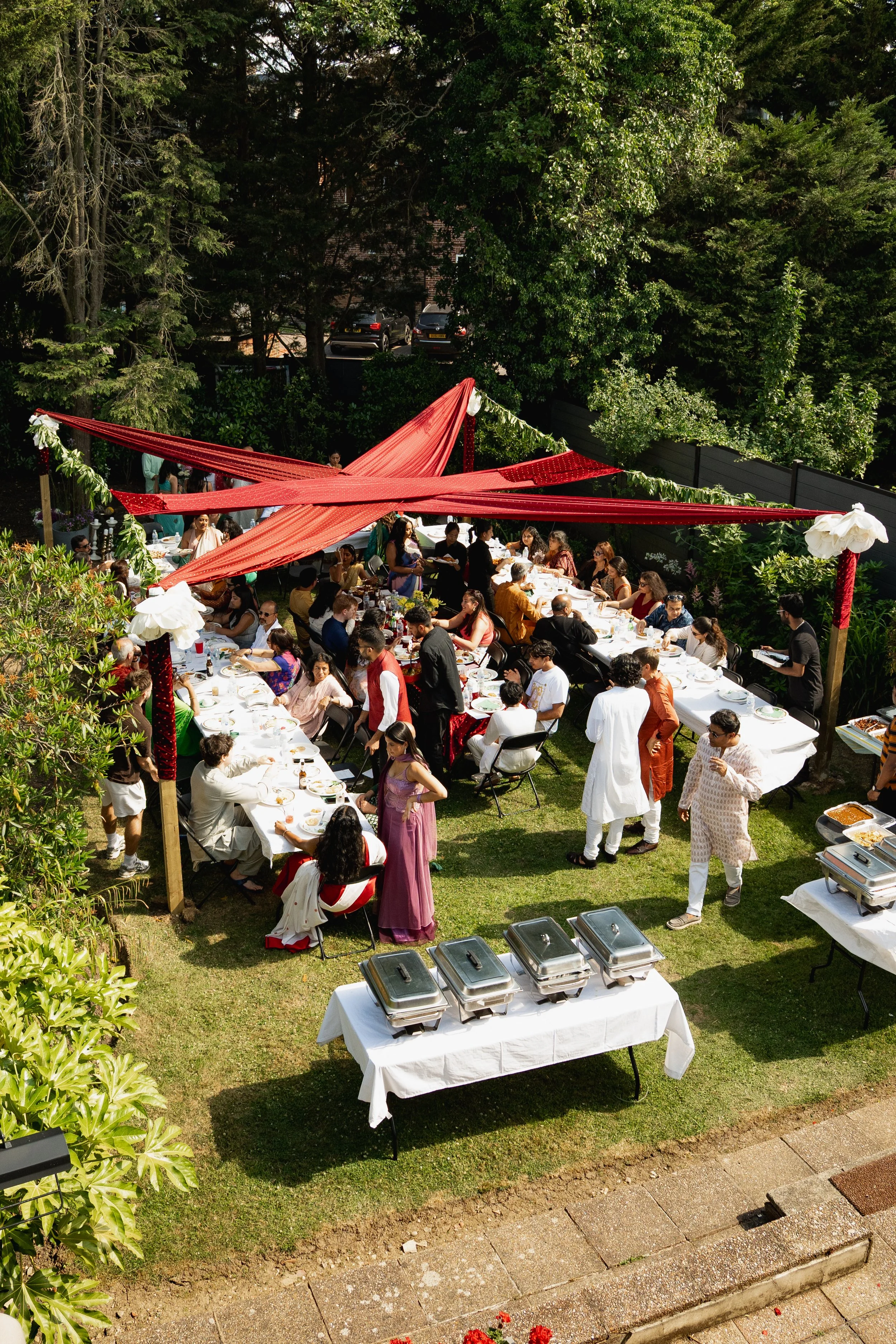 A large outdoor gathering with people seated at long tables under red fabric decorations, with buffet tables and lush green trees in the background.