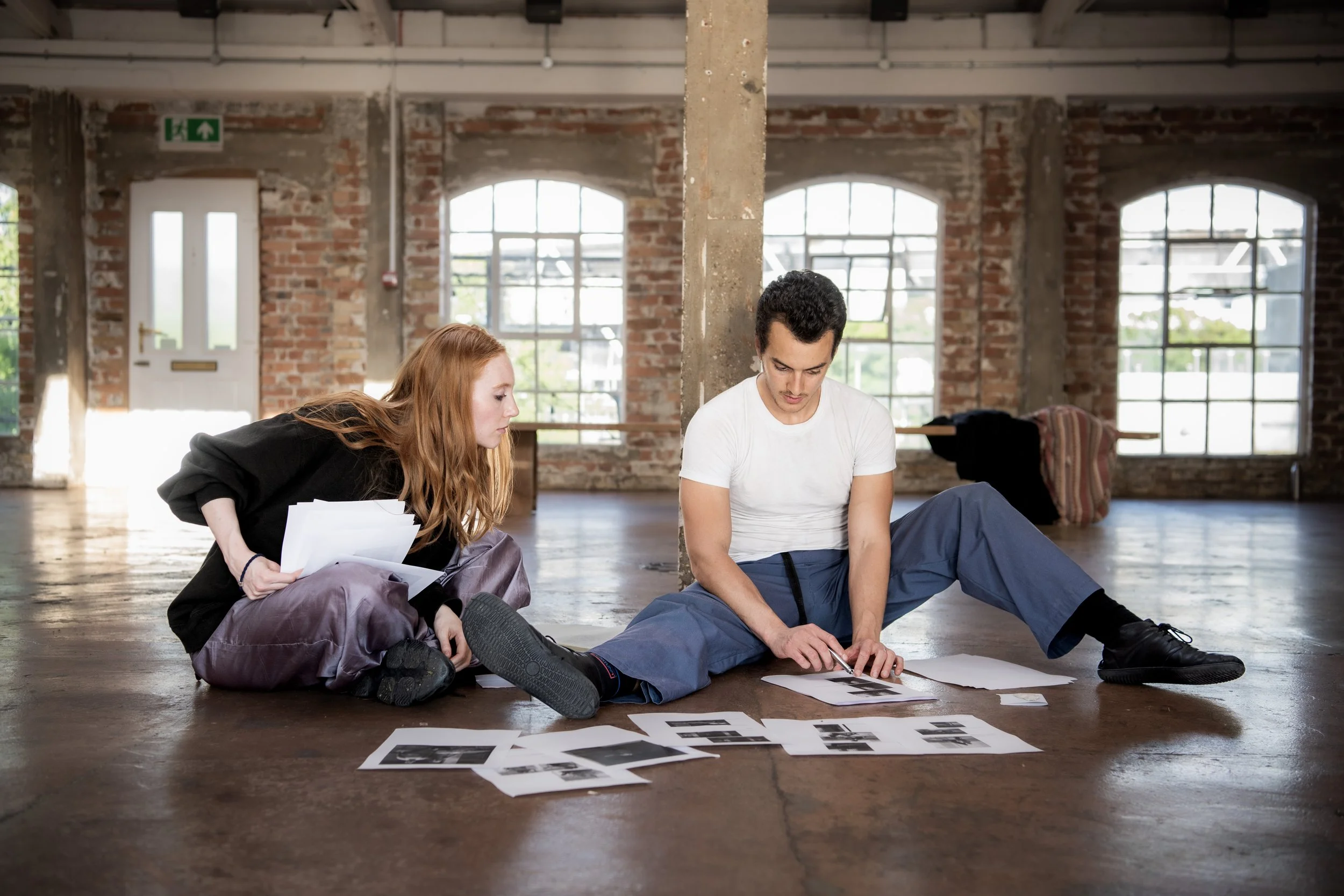 Two young people, a woman with red hair and a man with dark hair, sit on the floor of an industrial-style room with exposed brick walls and large windows. They are examining and discussing black-and-white photographs or documents spread on the floor.