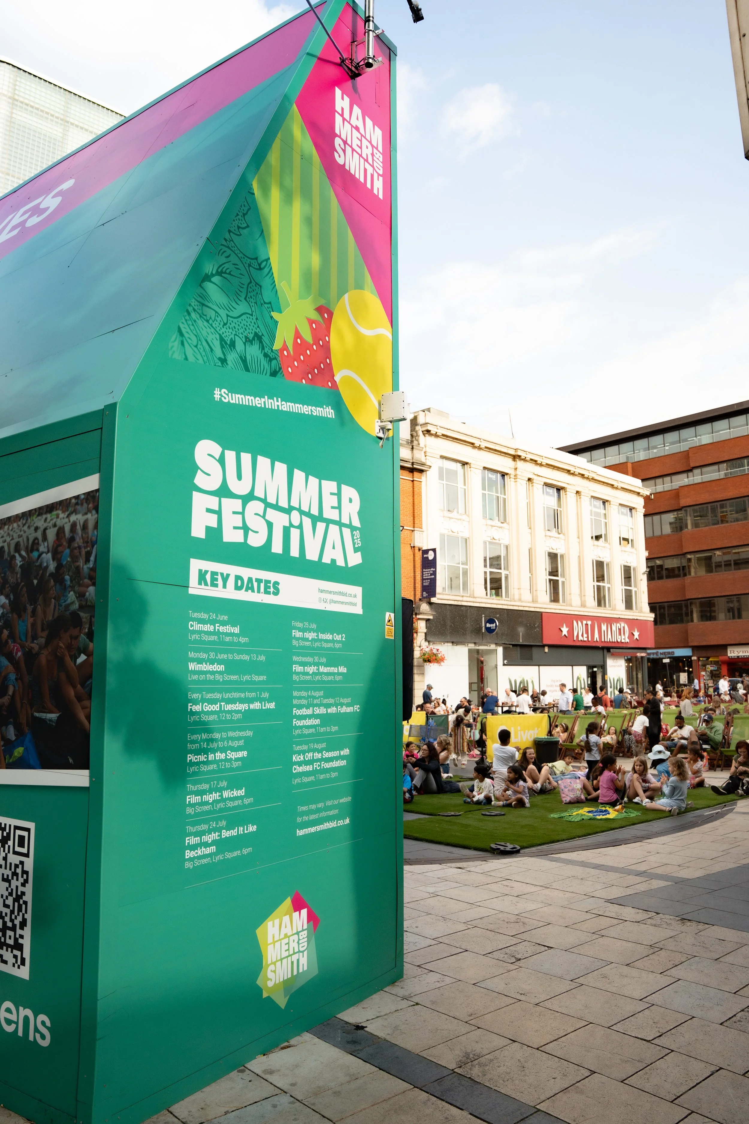 Large turquoise and pink sign at a summer festival listing key dates and events, with a crowd of people sitting and socializing on artificial grass in an urban plaza, and buildings in the background.