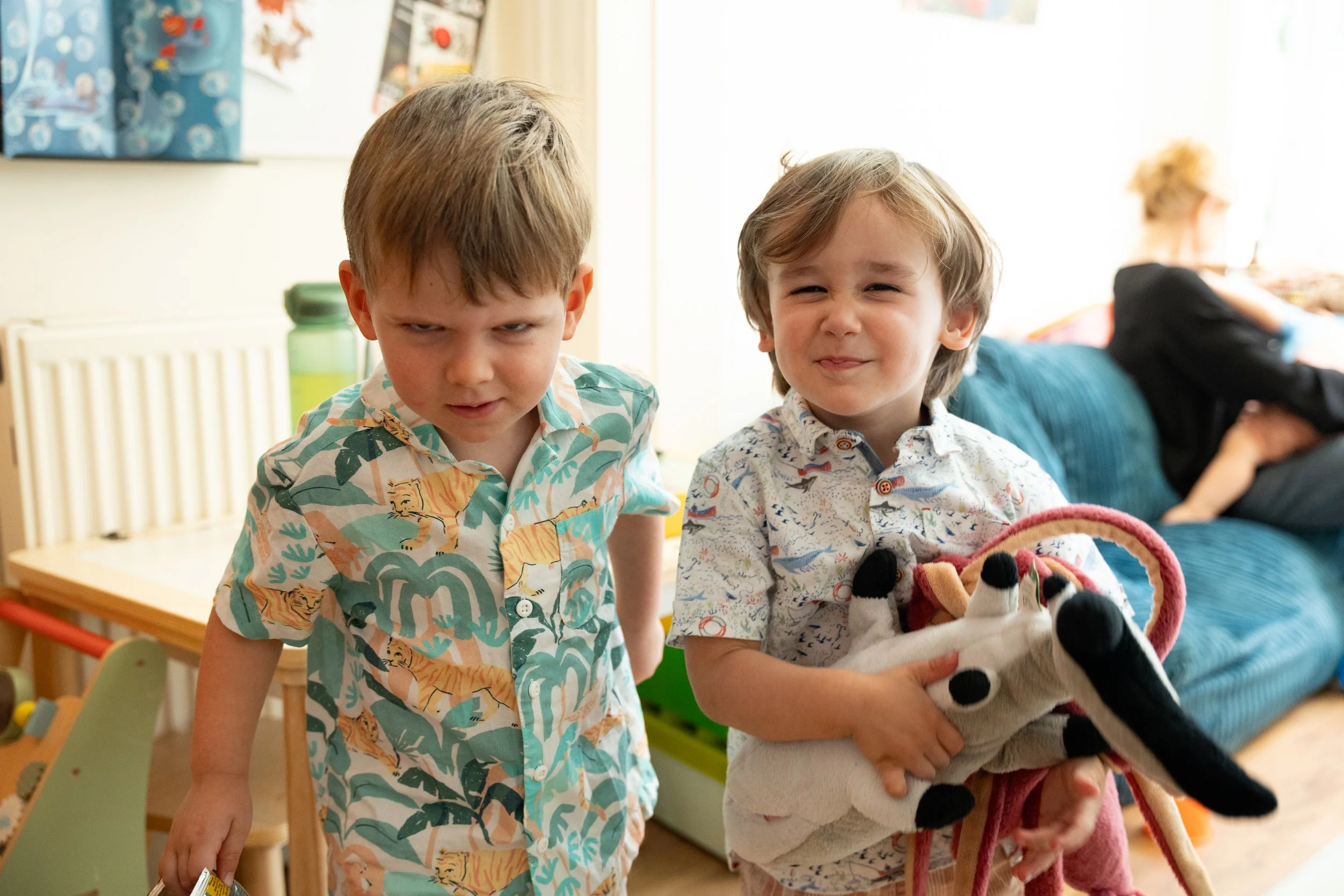 Two young boys with mischievous expressions standing indoors, with a woman lounging on a blue couch in the background.