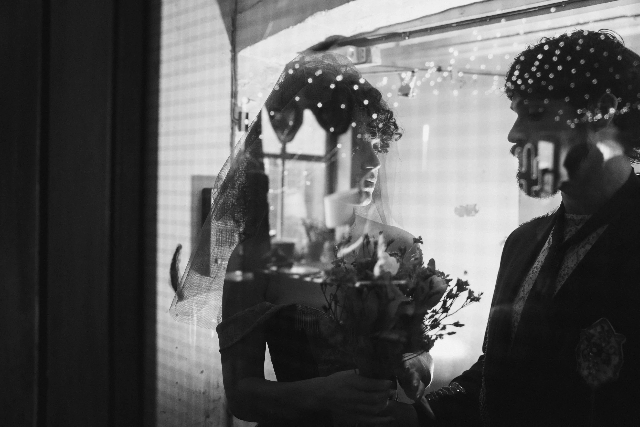 A black and white photo of a bride and a man in a police uniform looking at each other through a window with reflections and string lights.