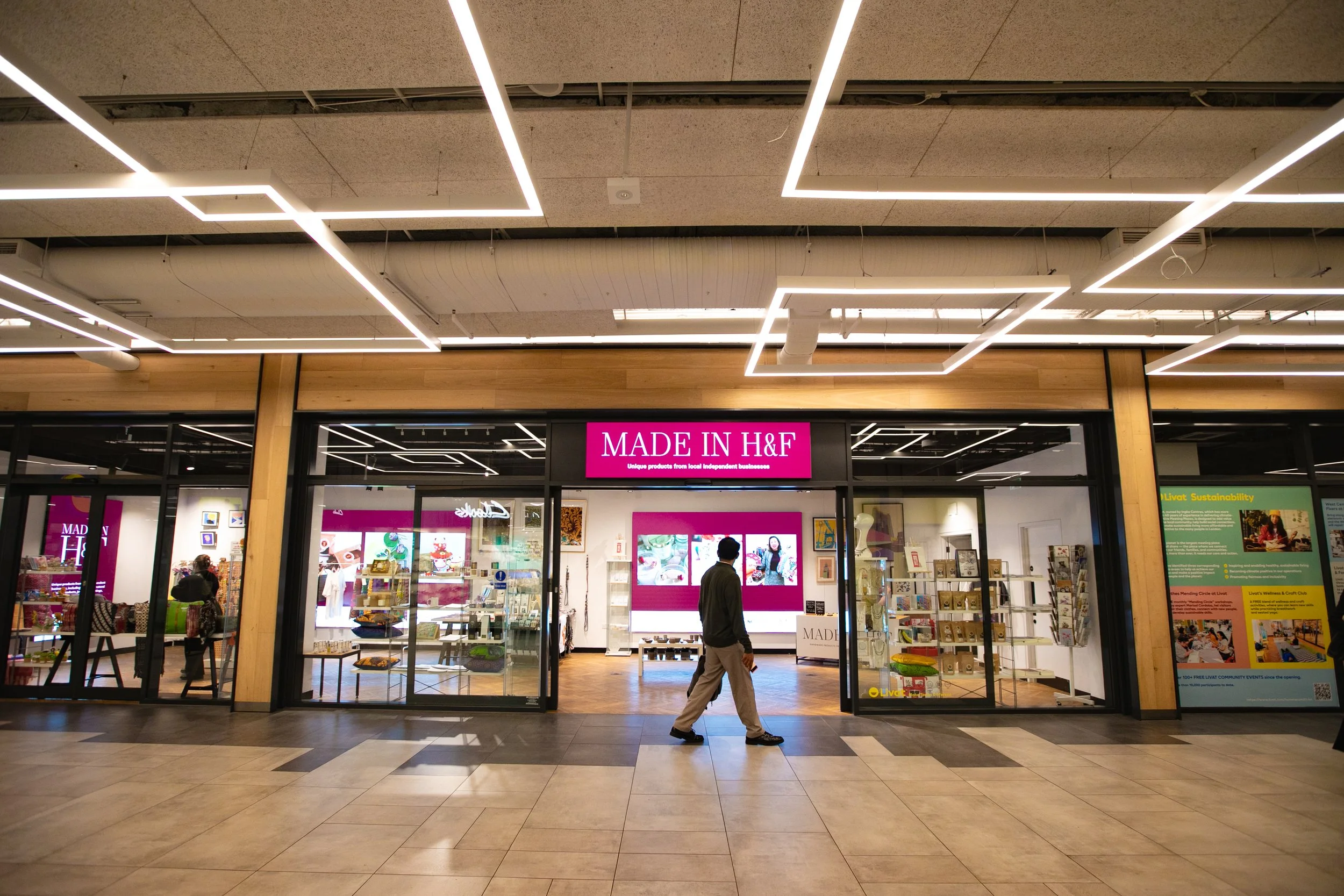 Man walking in front of a store called 'MADE IN H&F' in a shopping mall with a pink sign, glass storefront, and various products inside.