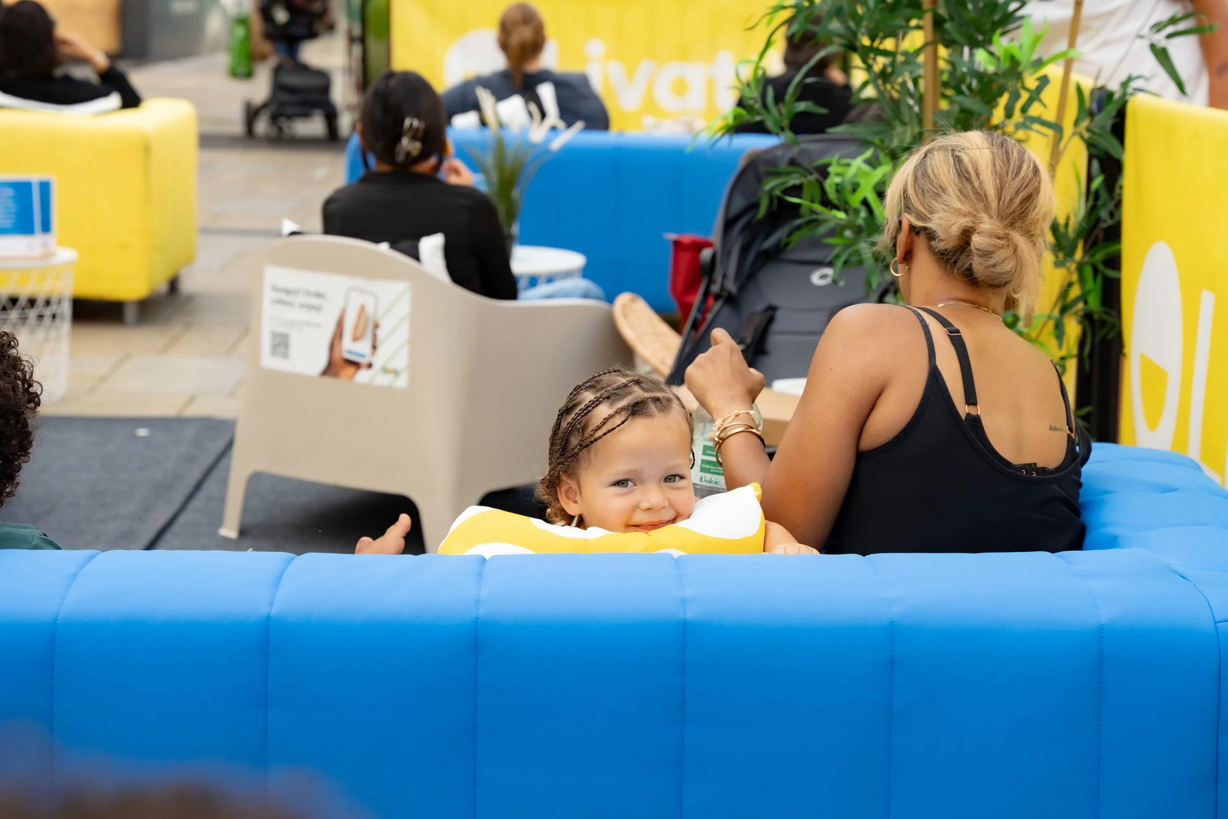 A young girl with braided hair smiling and resting her head on a yellow pillow inside a blue inflatable chair, with an adult woman sitting beside her in an indoor public space with colorful seating in the background.