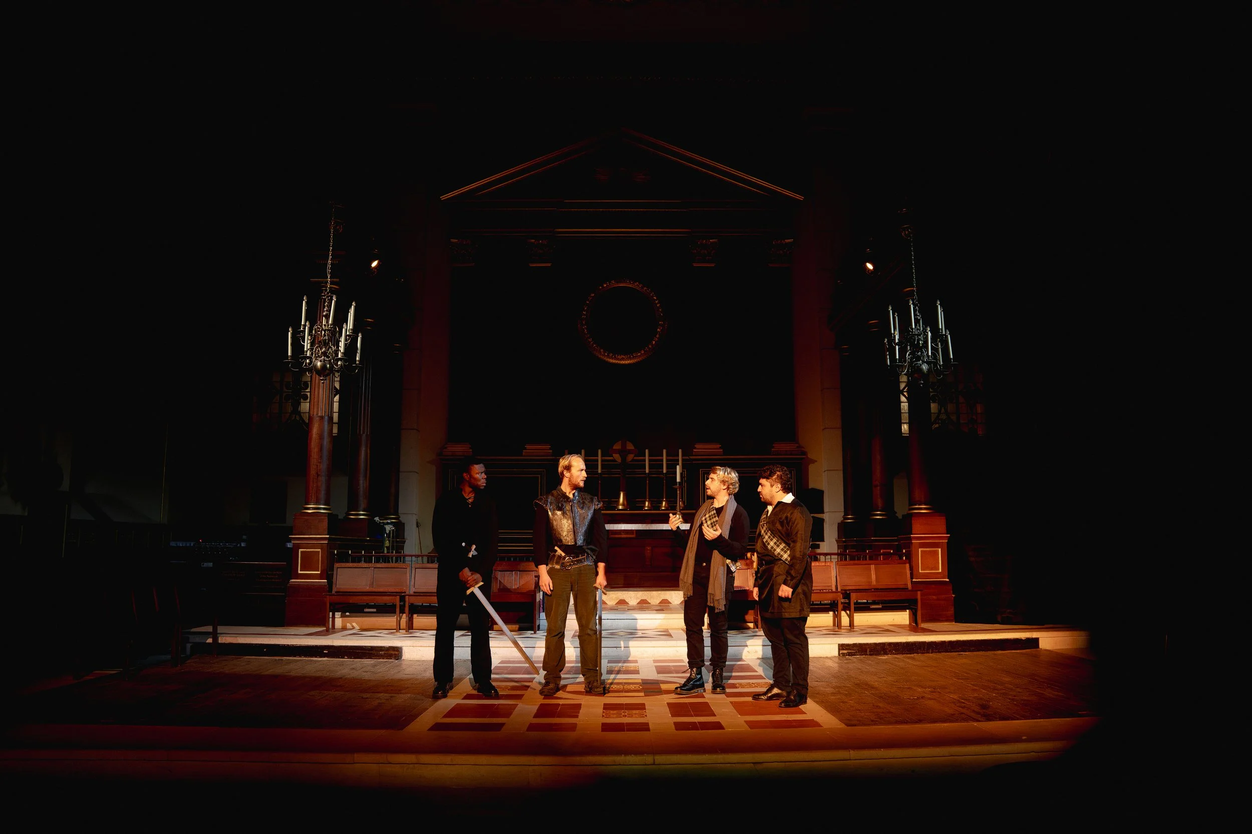 Four actors on stage in a theatrical performance, with a dark background and minimal lighting highlighting them, in a setting resembling an old courtroom or church.
