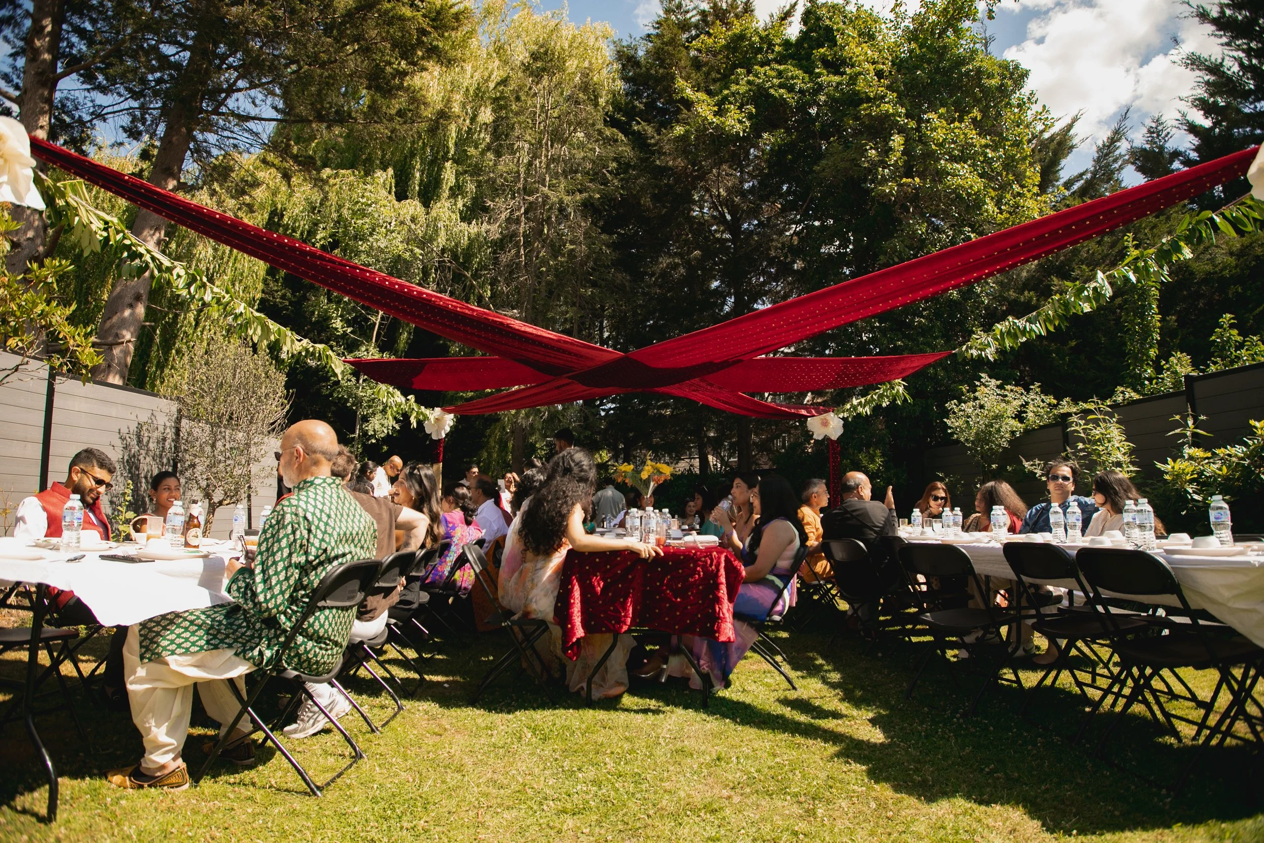 Outdoor gathering with people seated at long tables under red fabric drapes, surrounded by trees and greenery on a sunny day.
