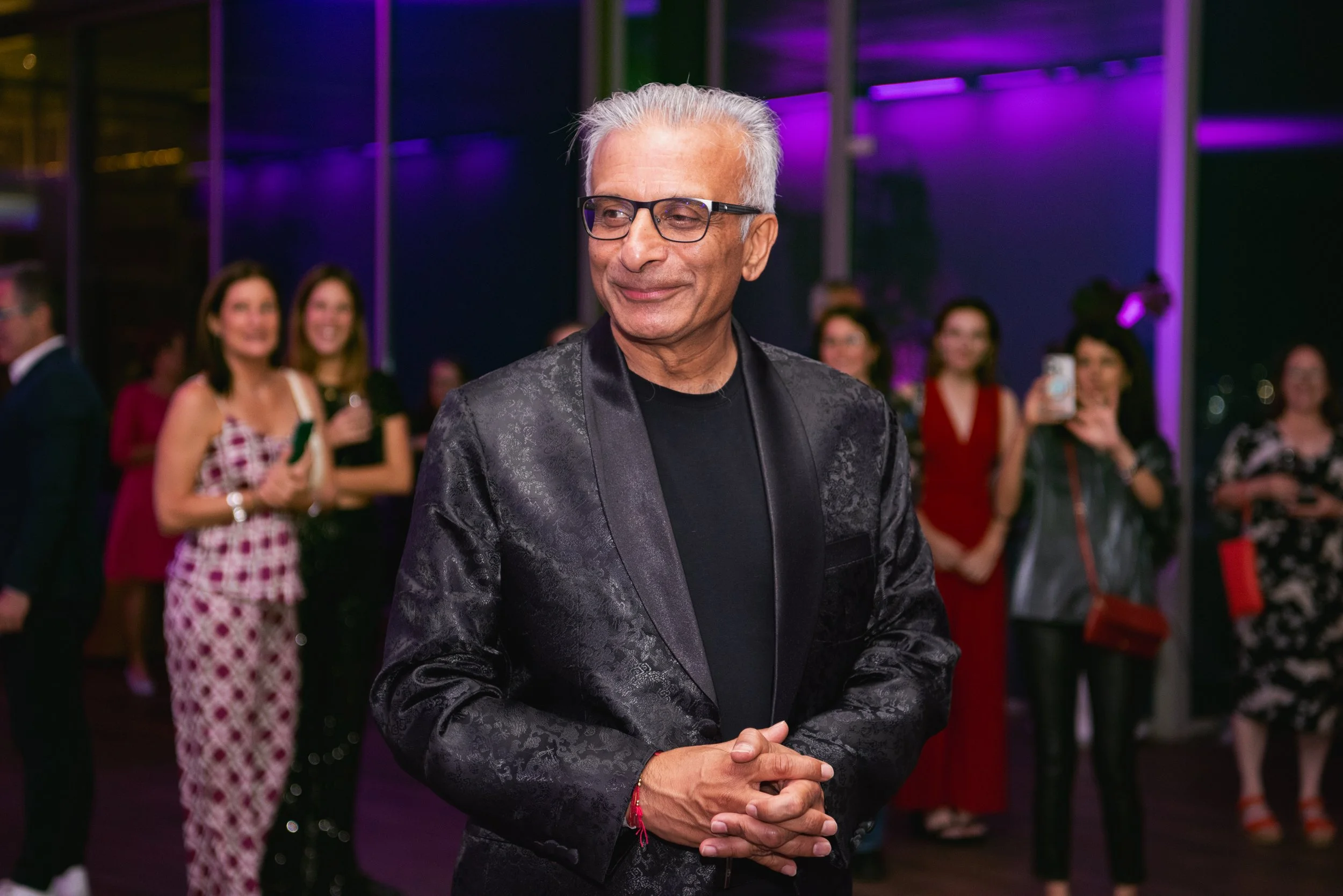 An older man with gray hair and glasses wearing a black patterned blazer and black shirt, standing with his hands clasped together at a social event with purple lighting. Several women in colorful dresses are in the background, some smiling and other