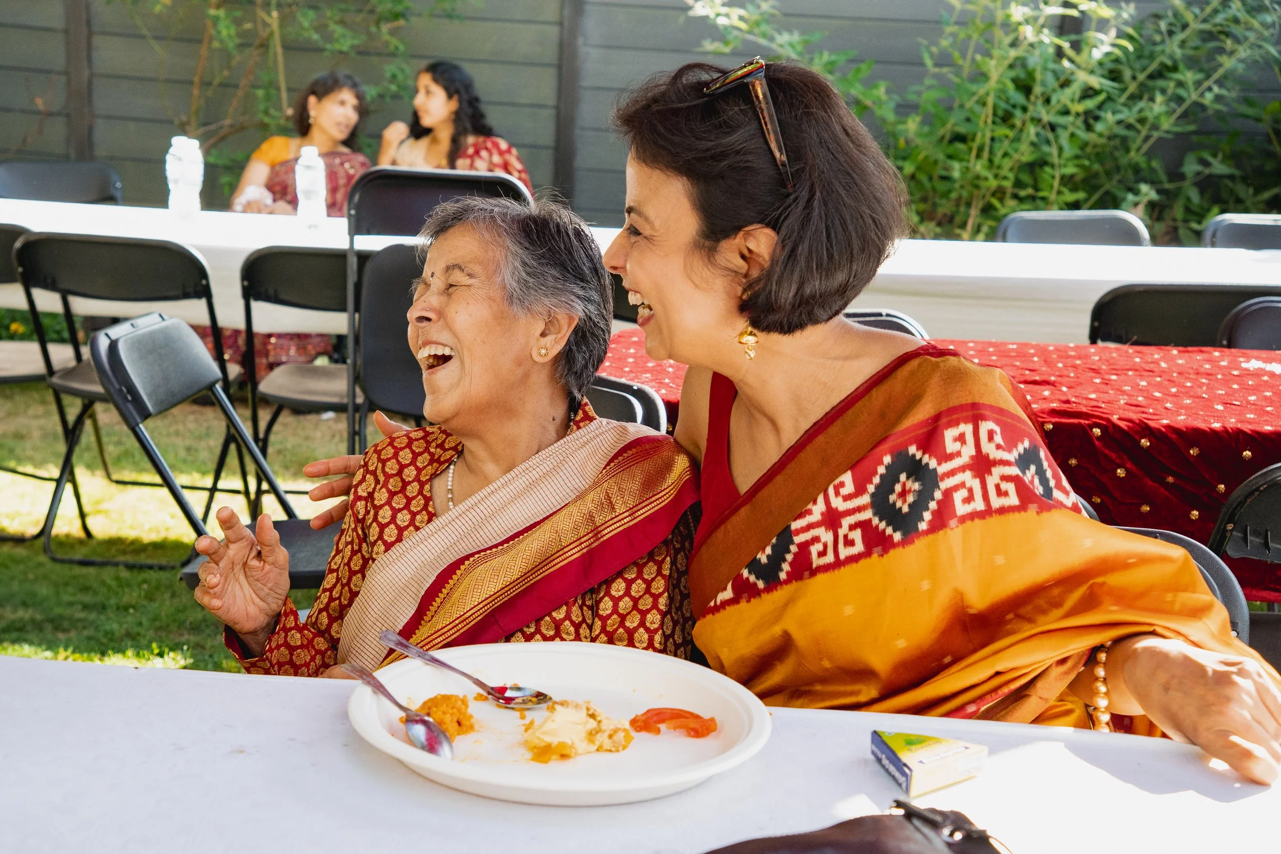 Two women, one elderly and one middle-aged, wearing traditional Indian sarees, are sitting at a table outdoors, laughing and enjoying a moment together. There are empty chairs and people in the background.