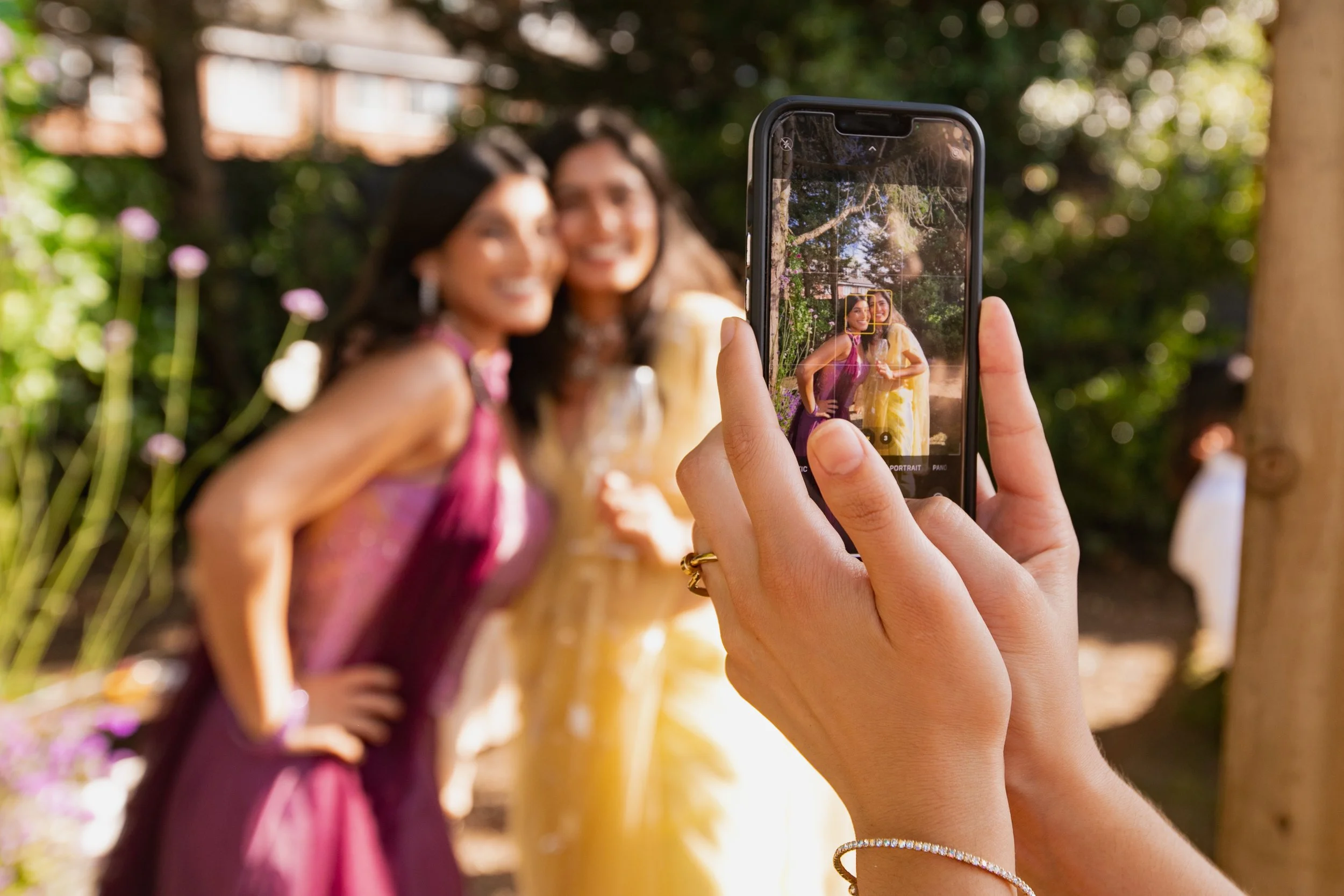 A person taking a photo with a smartphone of two women smiling and posing outside, with greenery and trees in the background.
