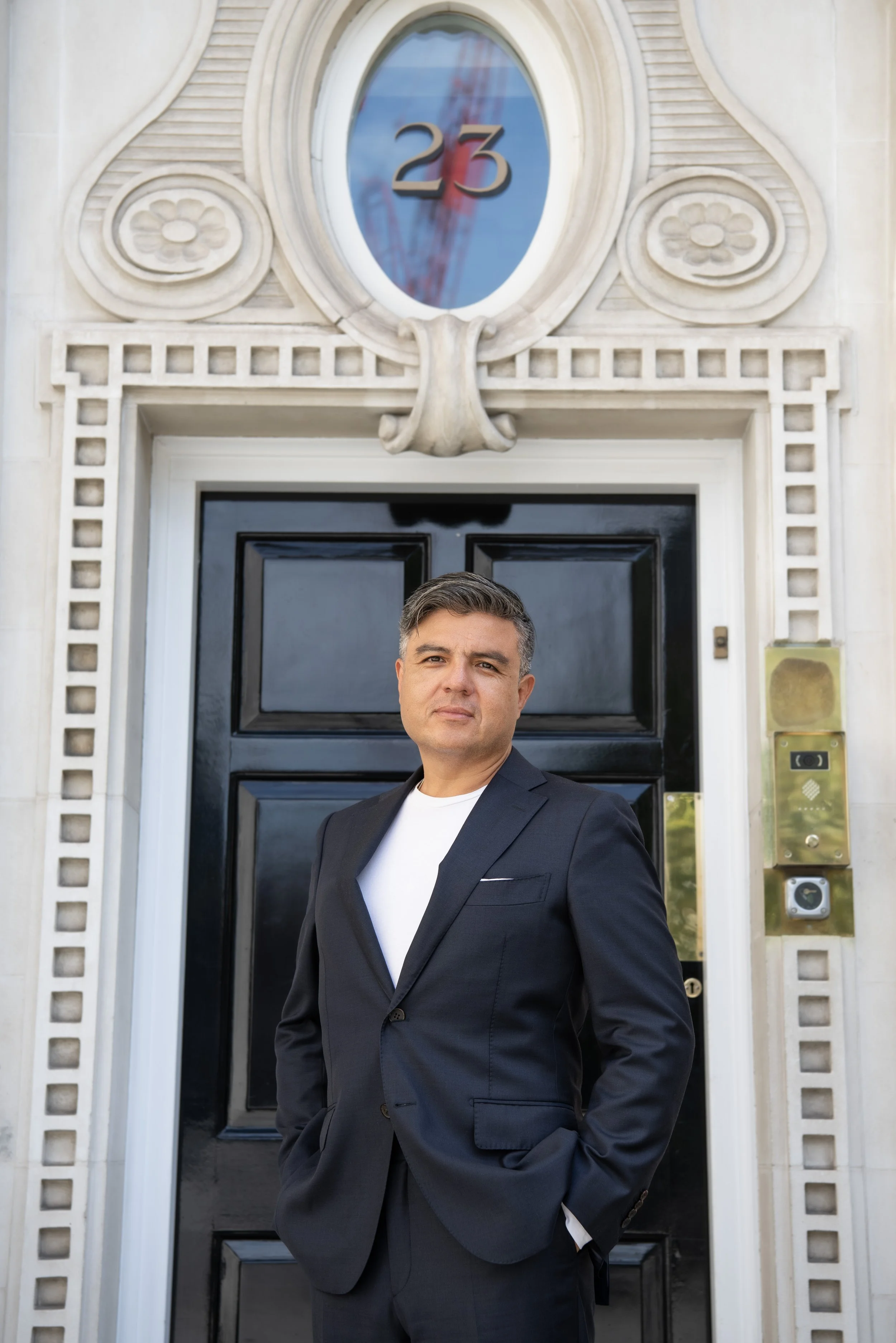 A man in a dark suit with a white shirt standing in front of a black door with the number 23 above it. The building has decorative white stonework around the door and a gold-colored intercom panel beside the door.