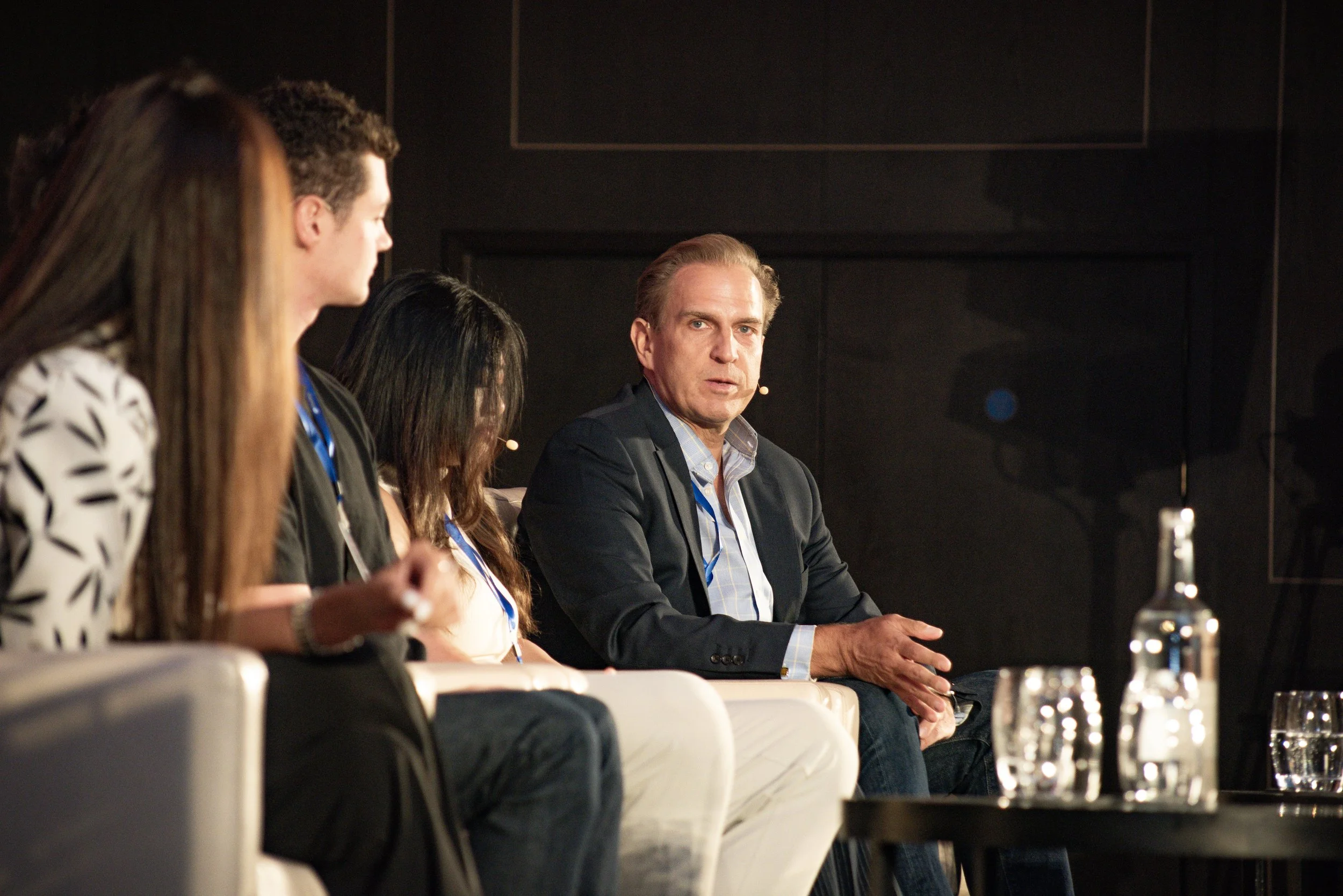 A panel of four people seated on stage during a discussion or conference, with dark background and bottles of water on a table in front of them.
