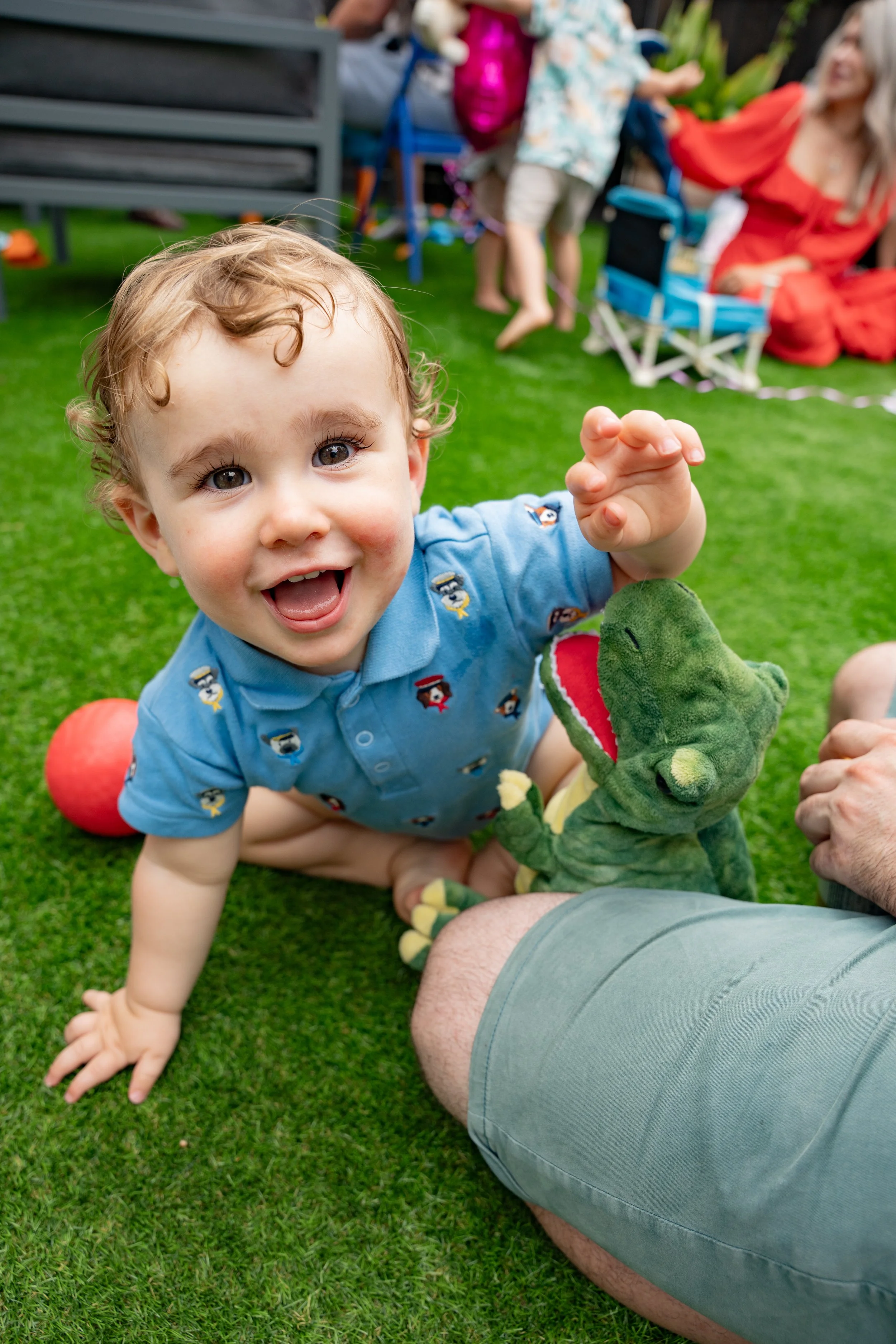 A young child with curly hair, wearing a blue shirt with colorful animal prints, is crawling on green grass and smiling at the camera while holding a green plush alligator toy. In the background, there are other children and an adult woman.