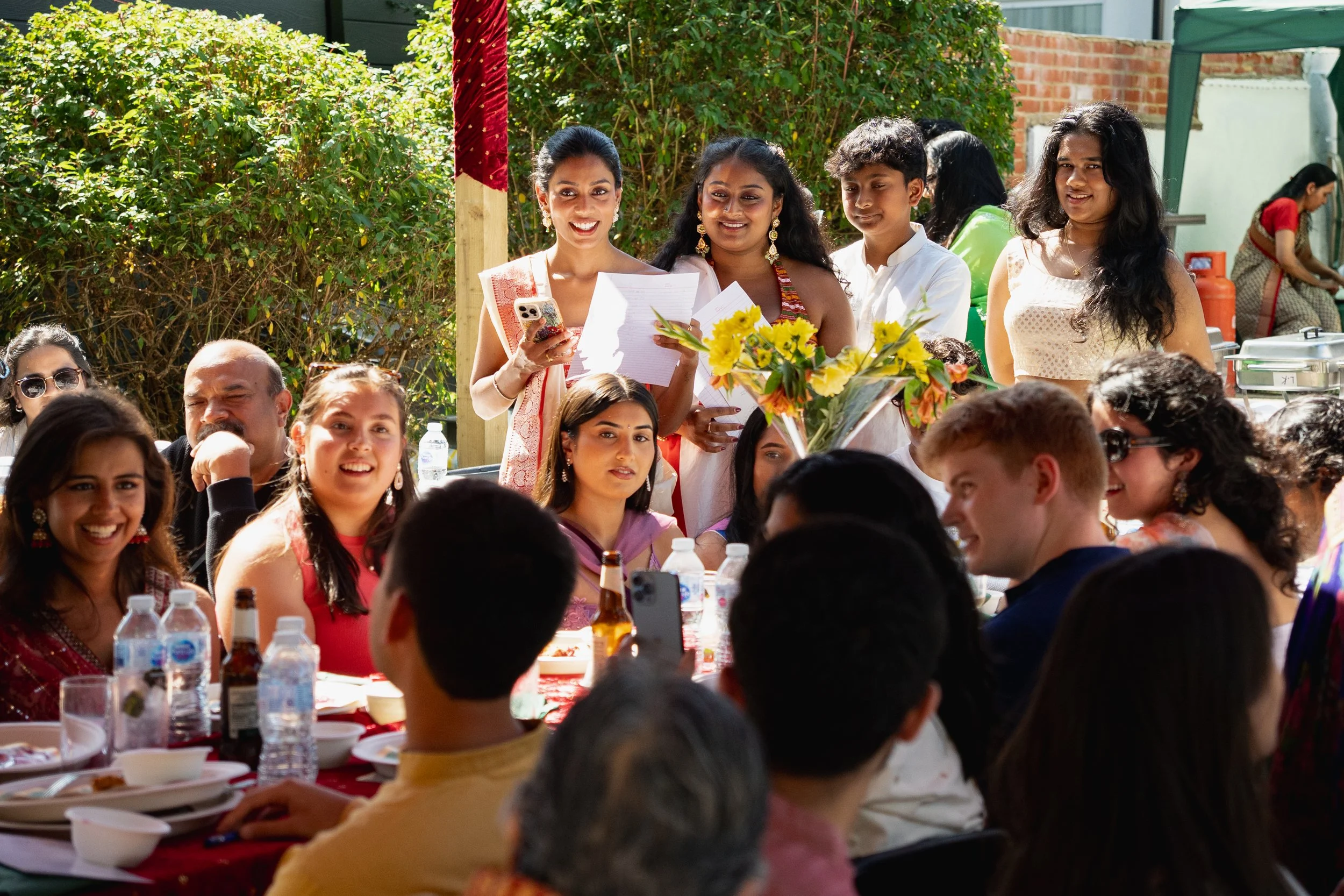 Wedding reception with people gathered at a table outdoors, women and men smiling, some holding paper, in festive attire, with a bouquet of yellow flowers on the table.