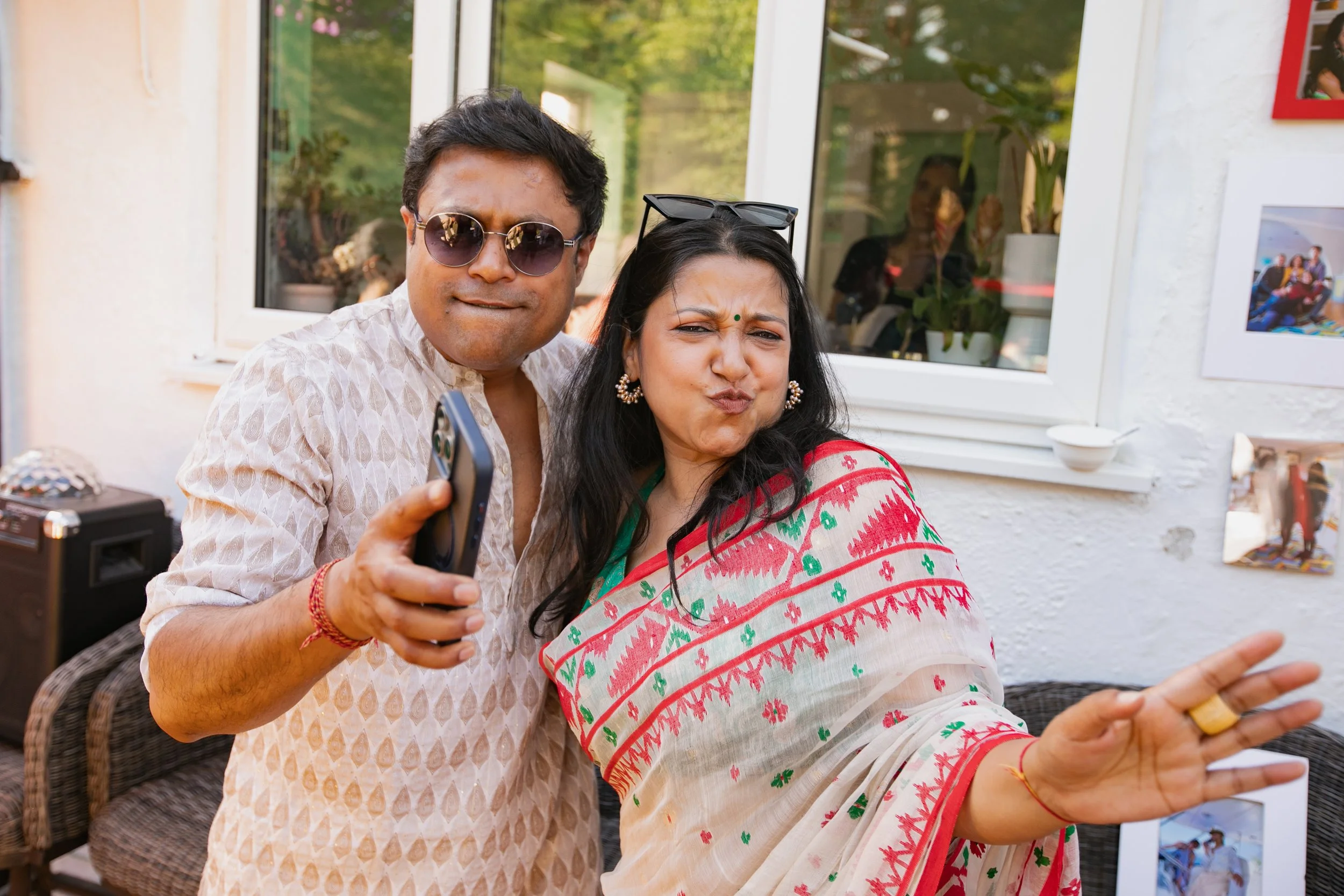 A man wearing sunglasses and a patterned shirt takes a selfie with a woman in traditional Indian attire, making a playful expression, in an outdoor setting with a house window and photos on the wall behind them.