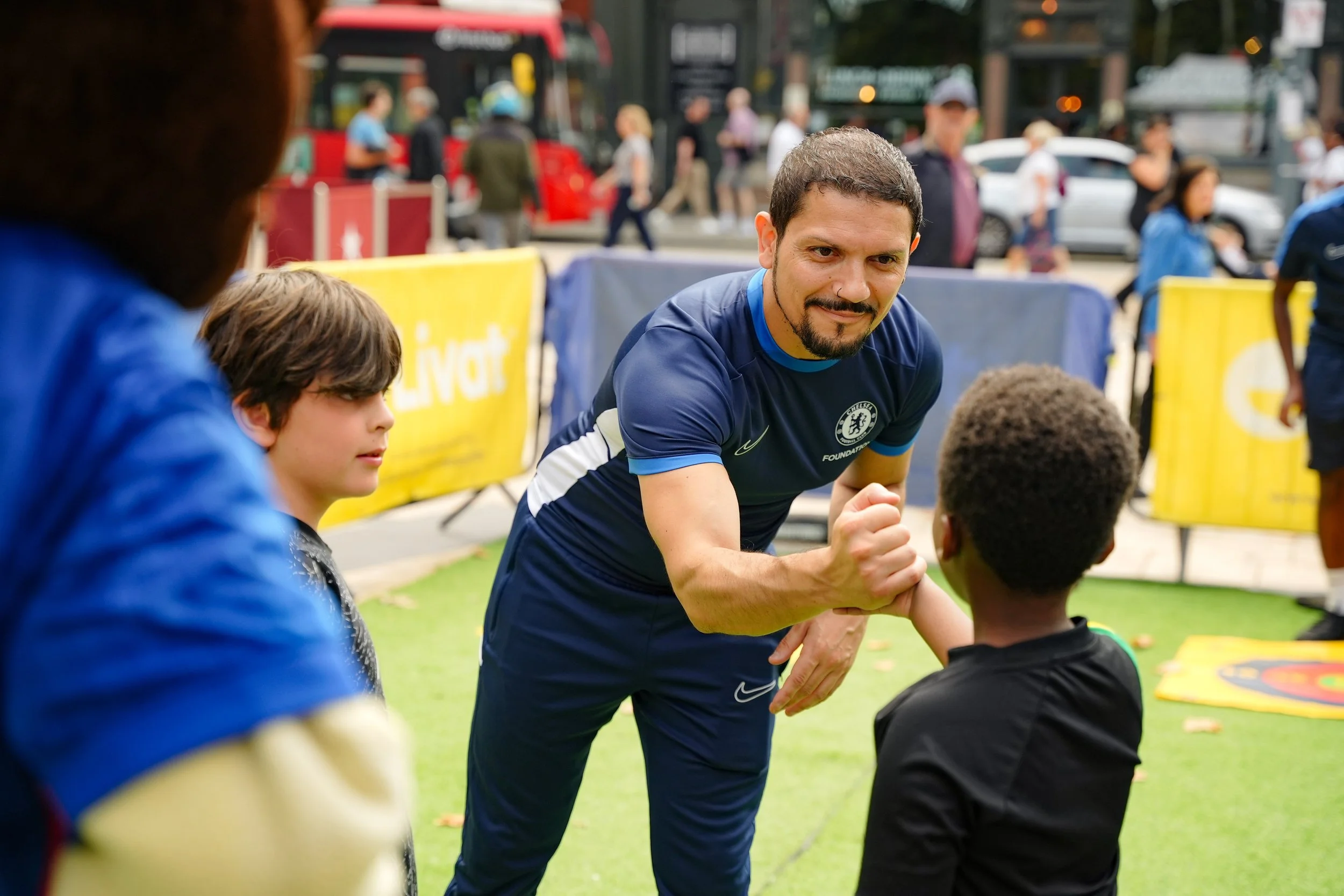 A man in a blue sports shirt greeting children outdoors, shaking hands and smiling, with people and vehicles blurred in the background.