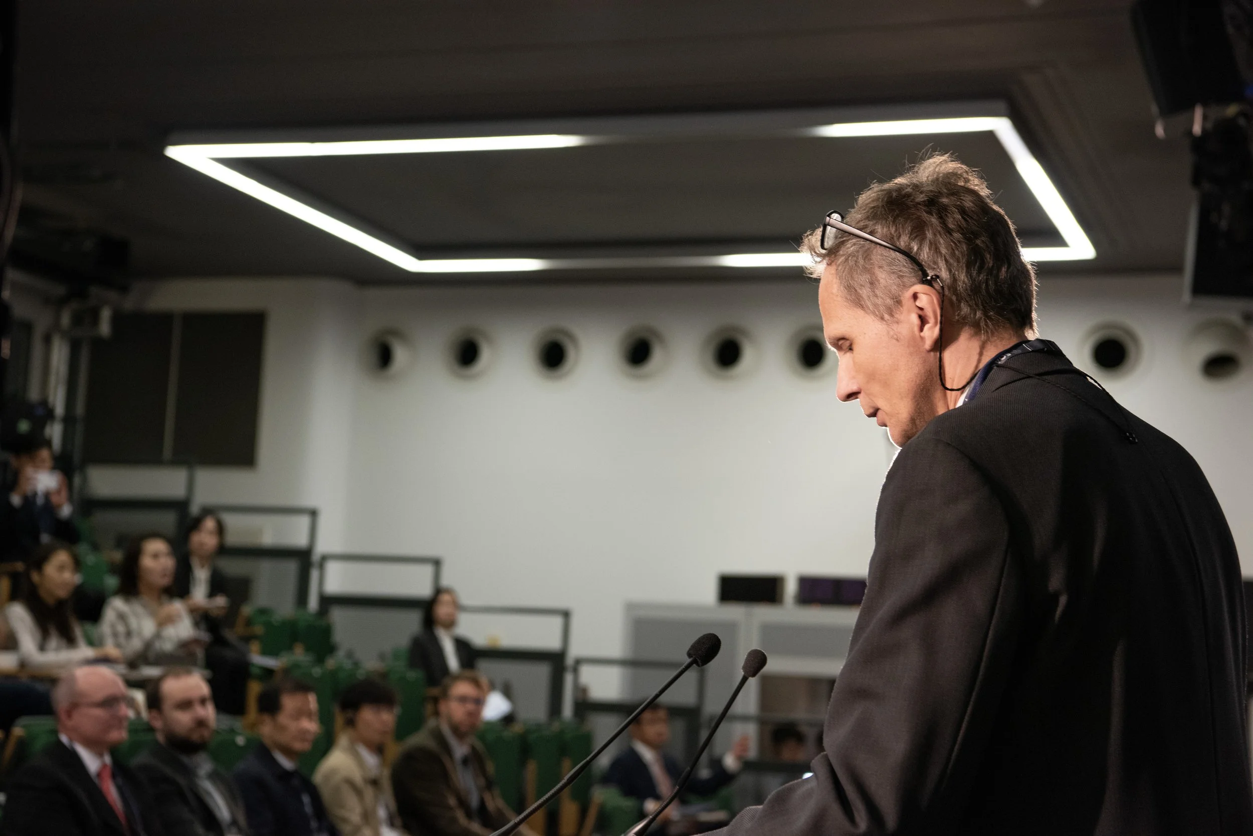 A man giving a speech at a podium during a conference or meeting, with an audience seated and listening attentively in a modern indoor venue.