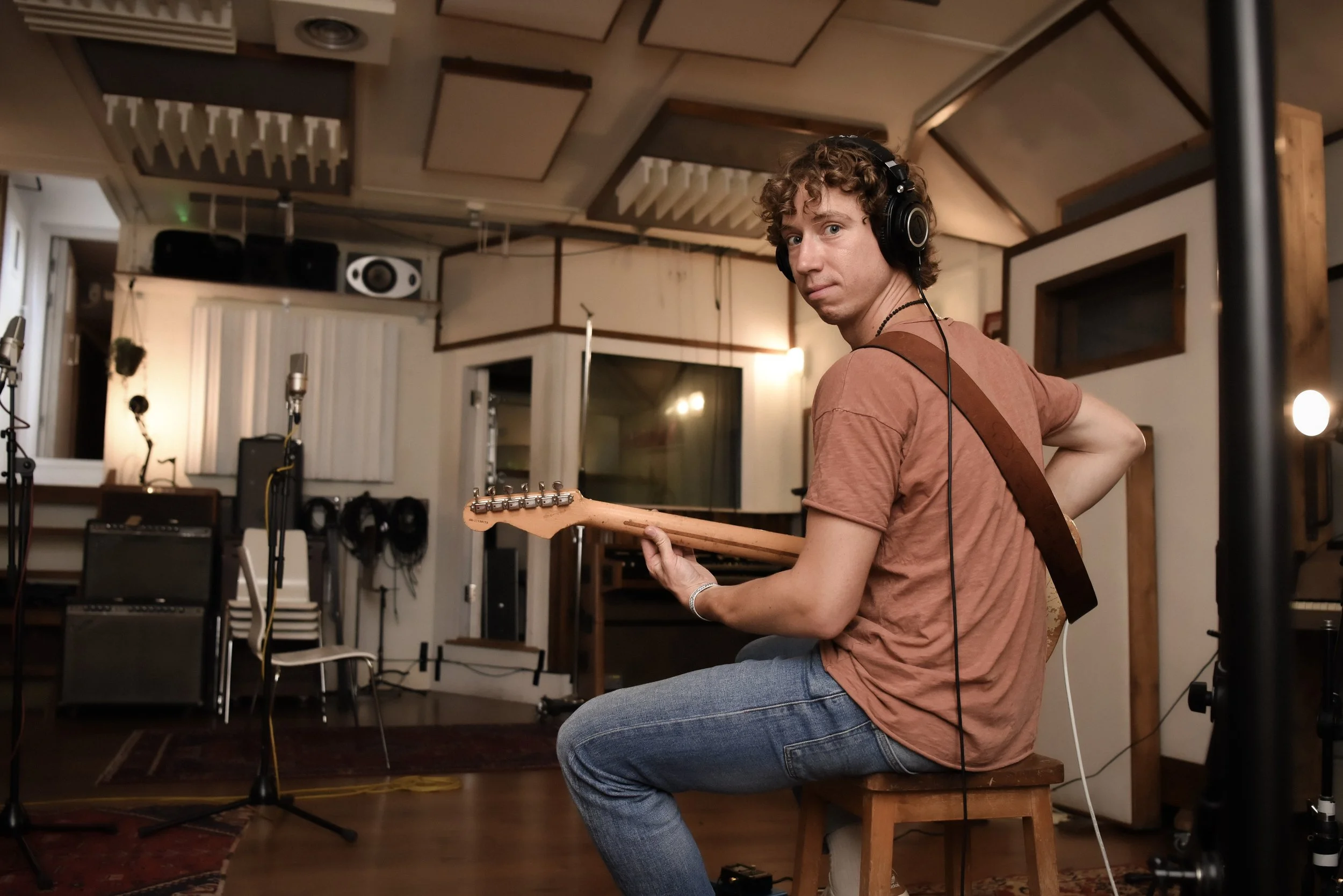 A young man with curly hair, wearing a brown t-shirt and jeans, sitting on a stool in a recording studio while playing an electric guitar. He is wearing large headphones and looking toward the camera.