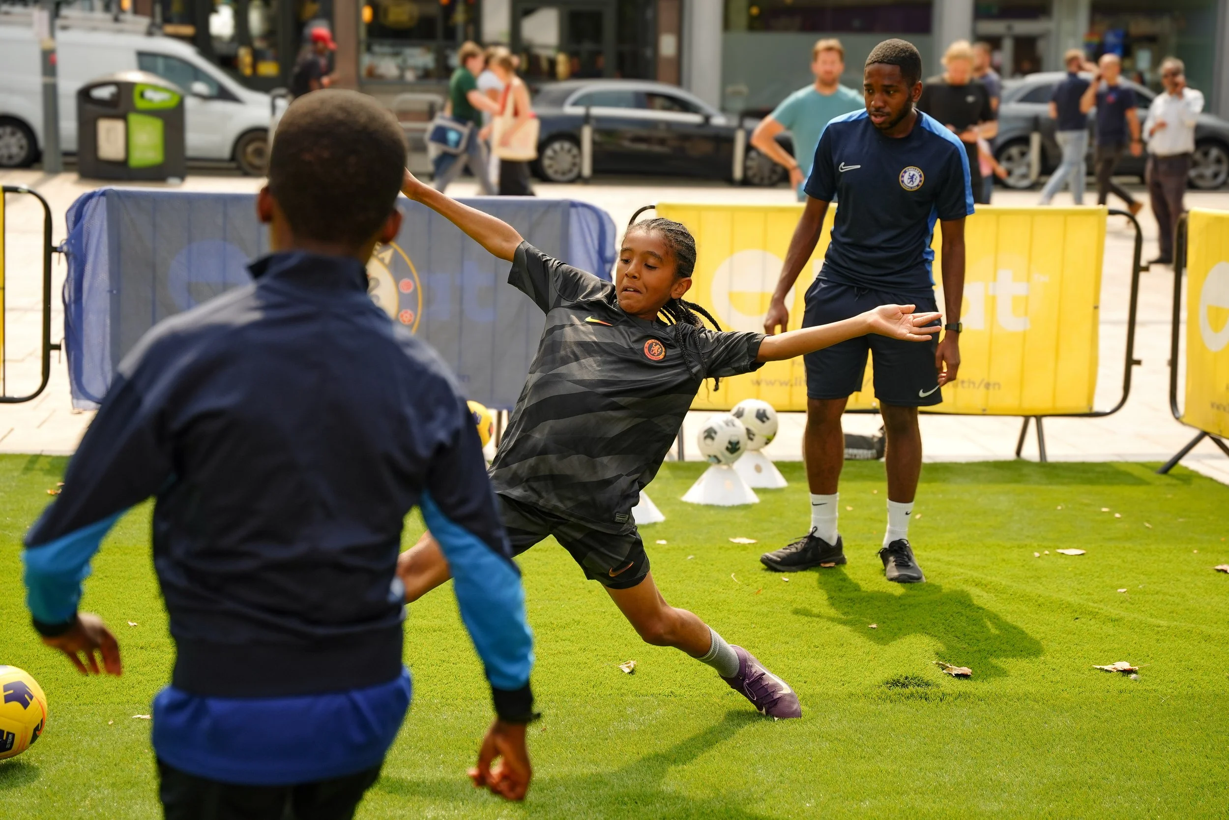 Two children playing soccer on a grassy field while a coach or referee watches, with several onlookers and cars in the background.