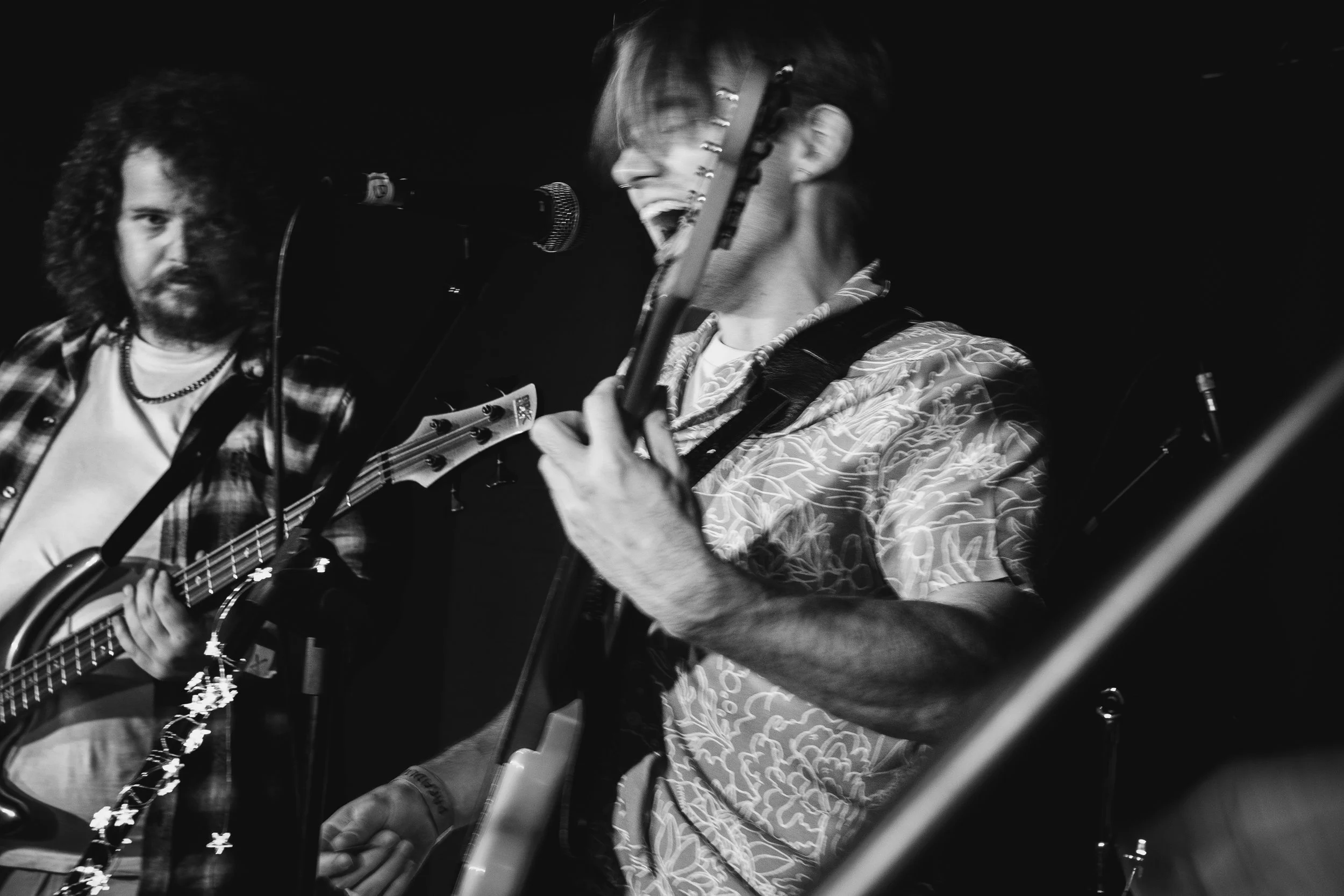 Two musicians performing on stage, one with an acoustic guitar and the other singing passionately into a microphone, both captured in black and white.