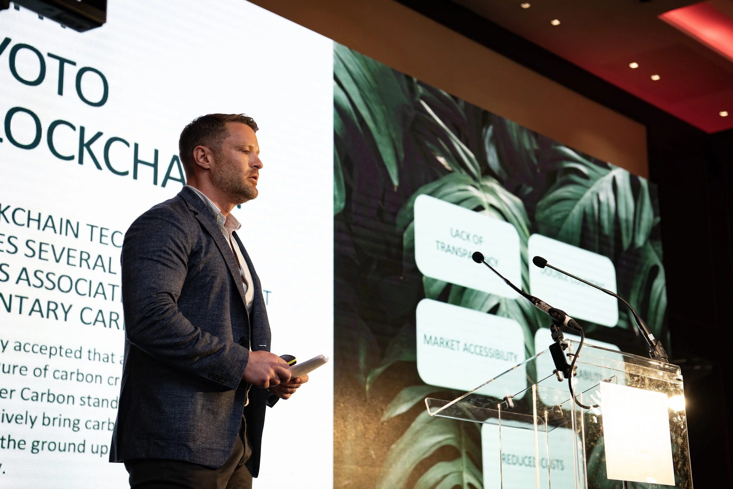 Man giving a presentation on stage at a conference or seminar, with a large screen behind him displaying text and graphics.