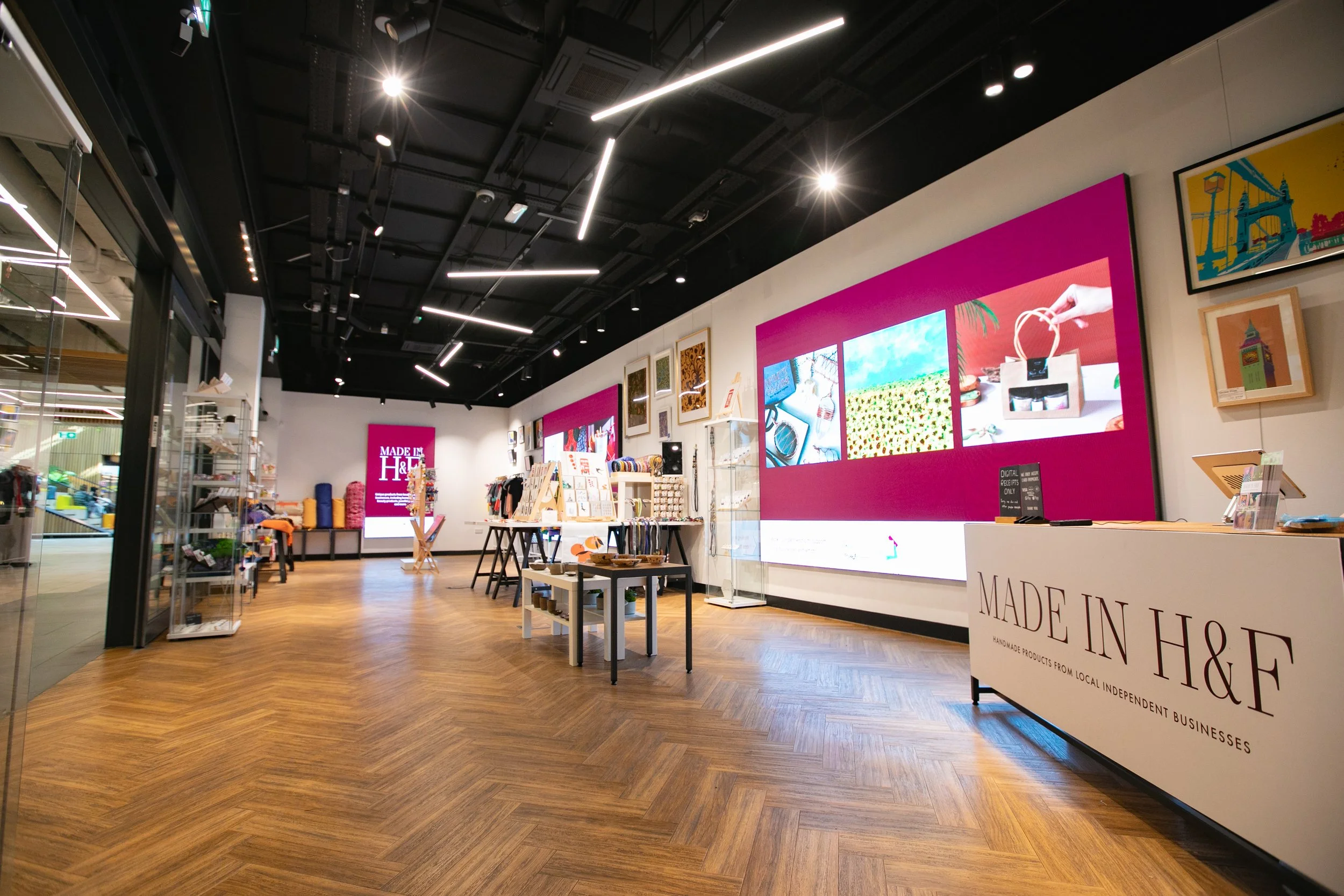 Inside a retail store with wooden flooring and modern lighting. Displays of handmade goods and framed artwork are arranged around. A large digital screen on the wall shows colorful images. A counter with a sign reads 'Made in H & F: Handmade products