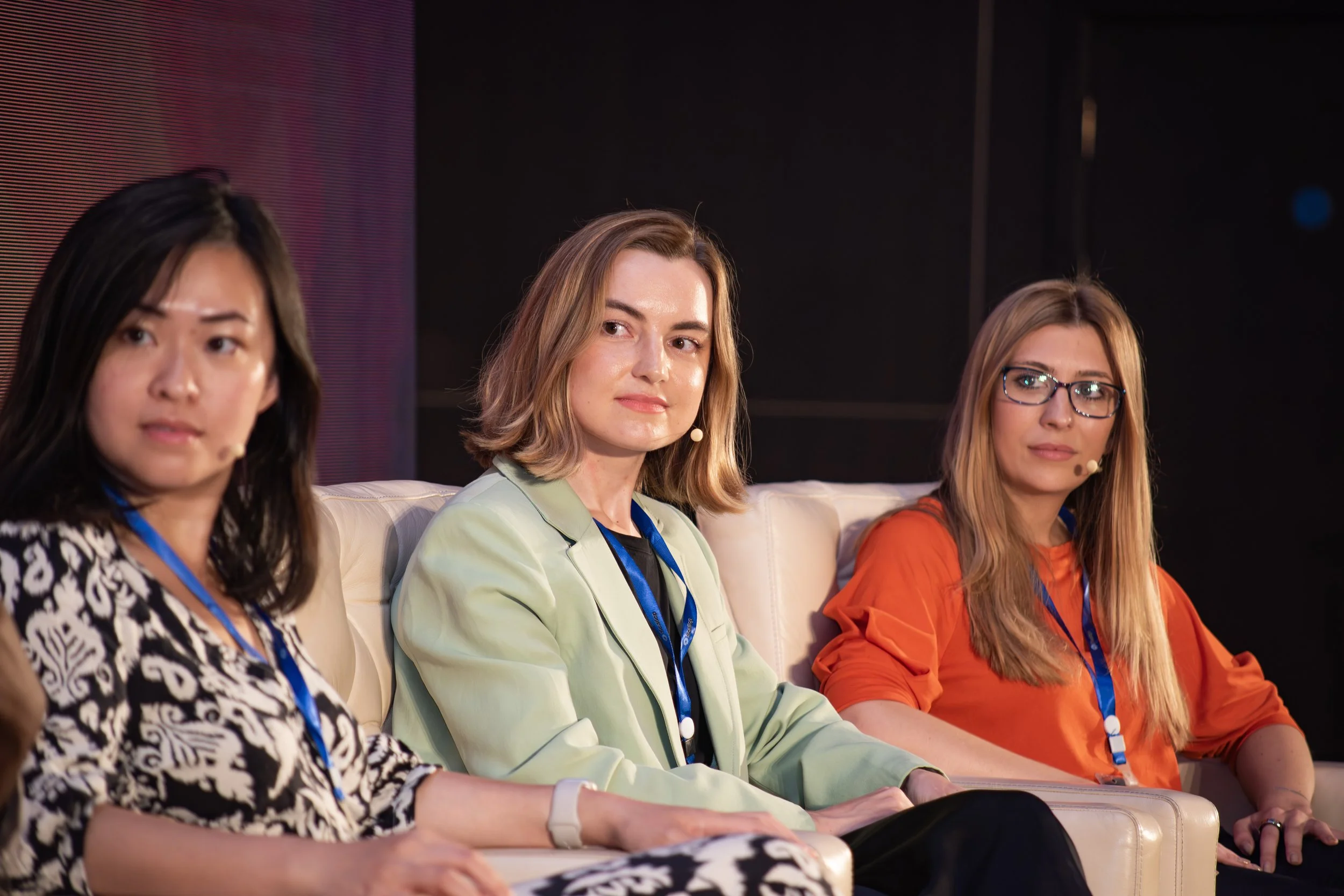 Three women sitting on a white sofa during a panel discussion or conference, each wearing a lanyard and microphone headset, with a dark background.