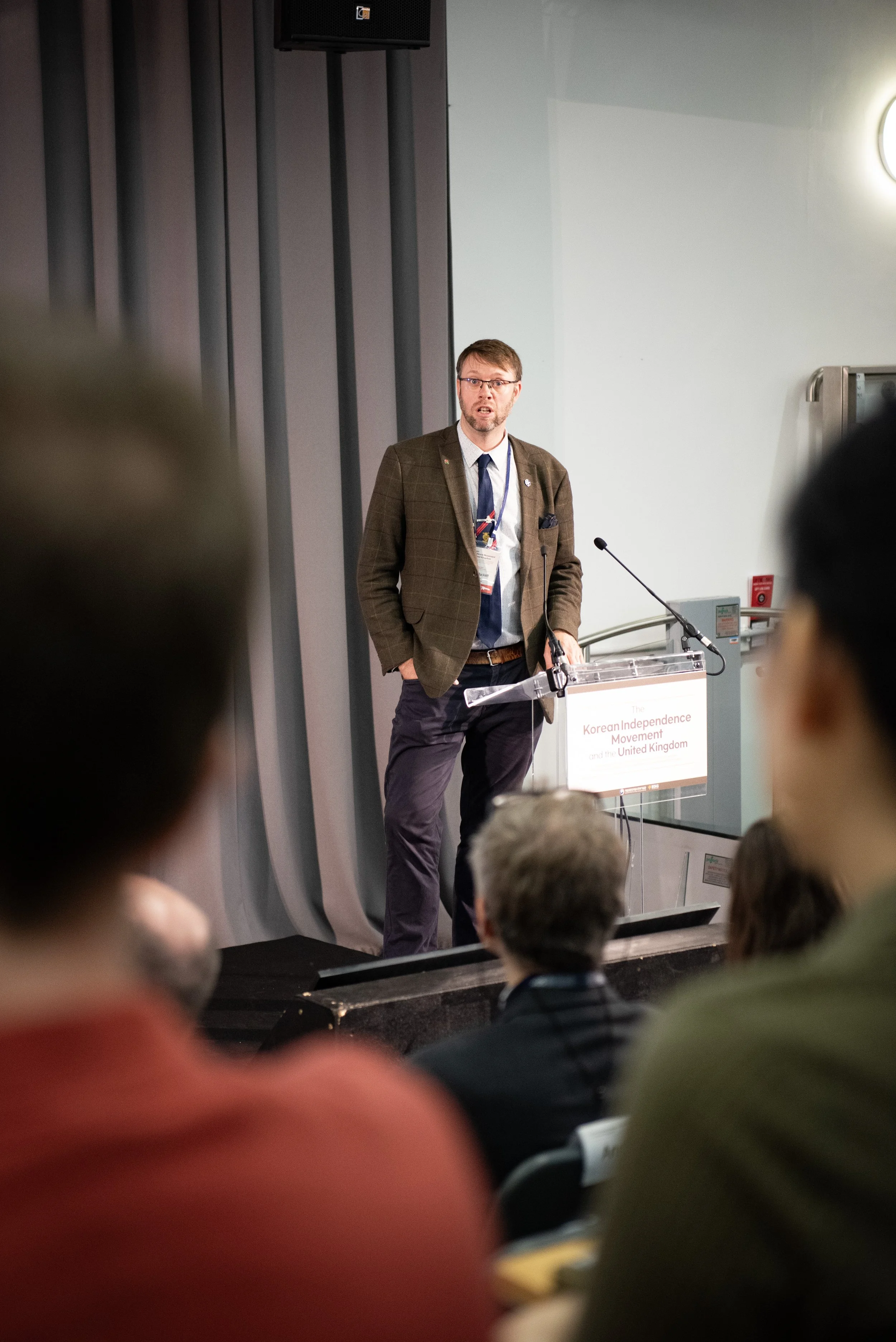 A man standing at a podium giving a lecture to an audience, with a sign on the podium reading 'The Korean Independence Movement and the United Kingdom.'