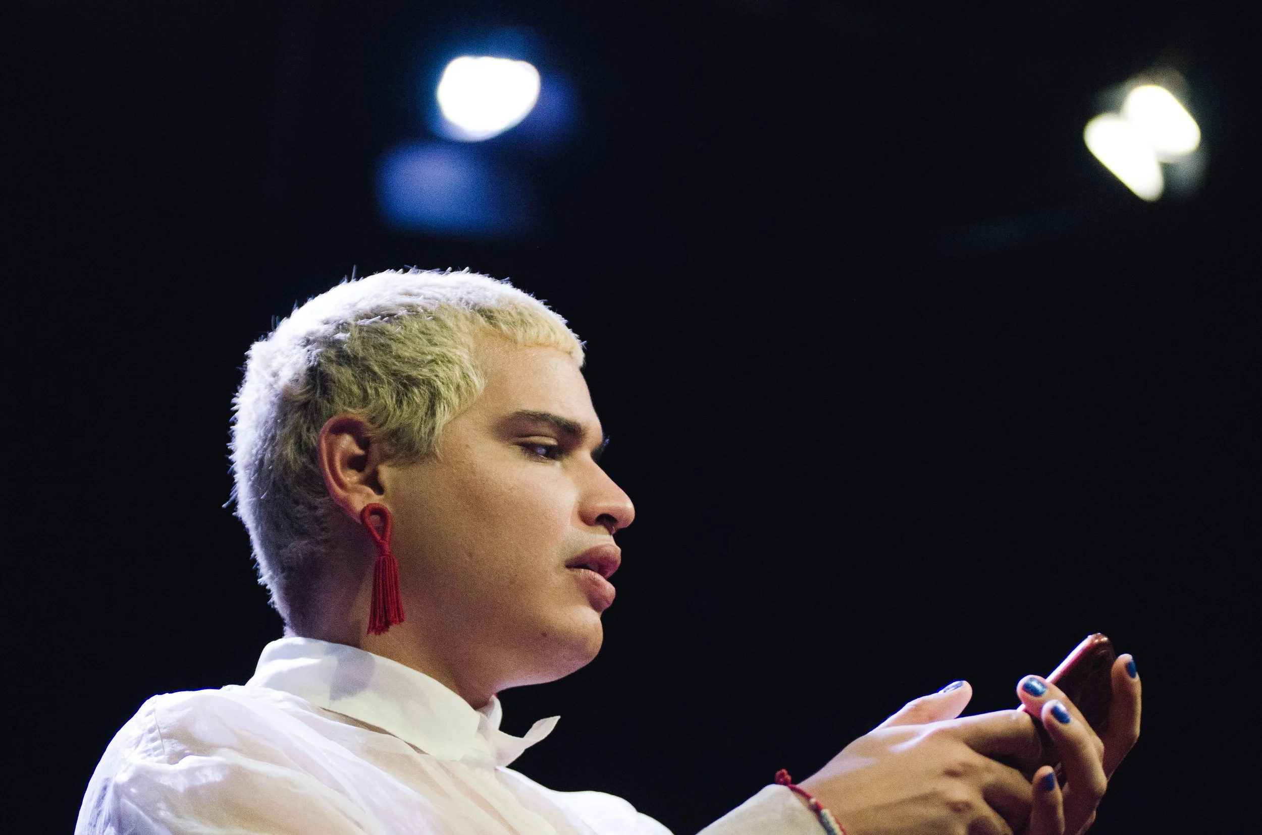 A woman with short bleached hair wearing red earrings and a white shirt, looking at her phone against a dark background with bright lights.