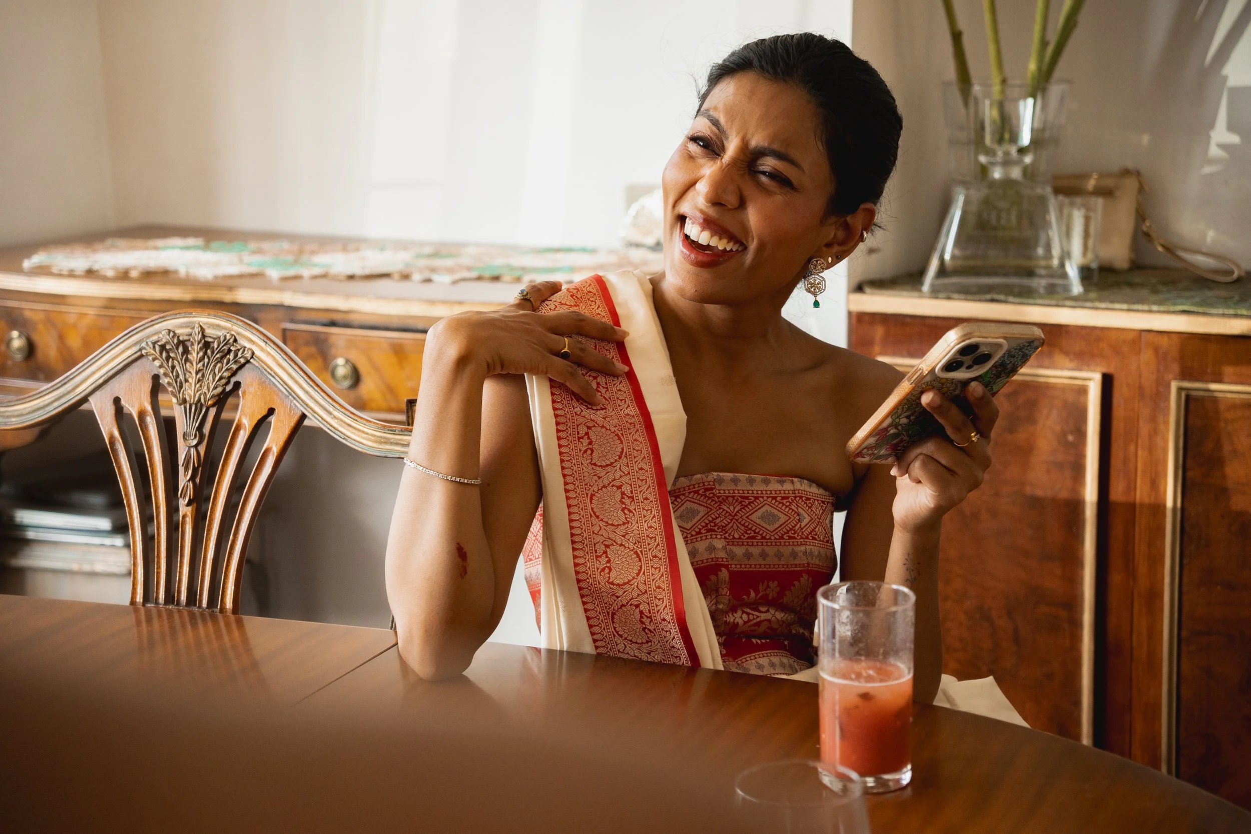 Woman with short dark hair, wearing traditional attire, sitting at a wooden dining table, smiling, holding a phone in her right hand, with a glass of pink beverage in front of her, and a wooden sideboard with decorative items in the background.