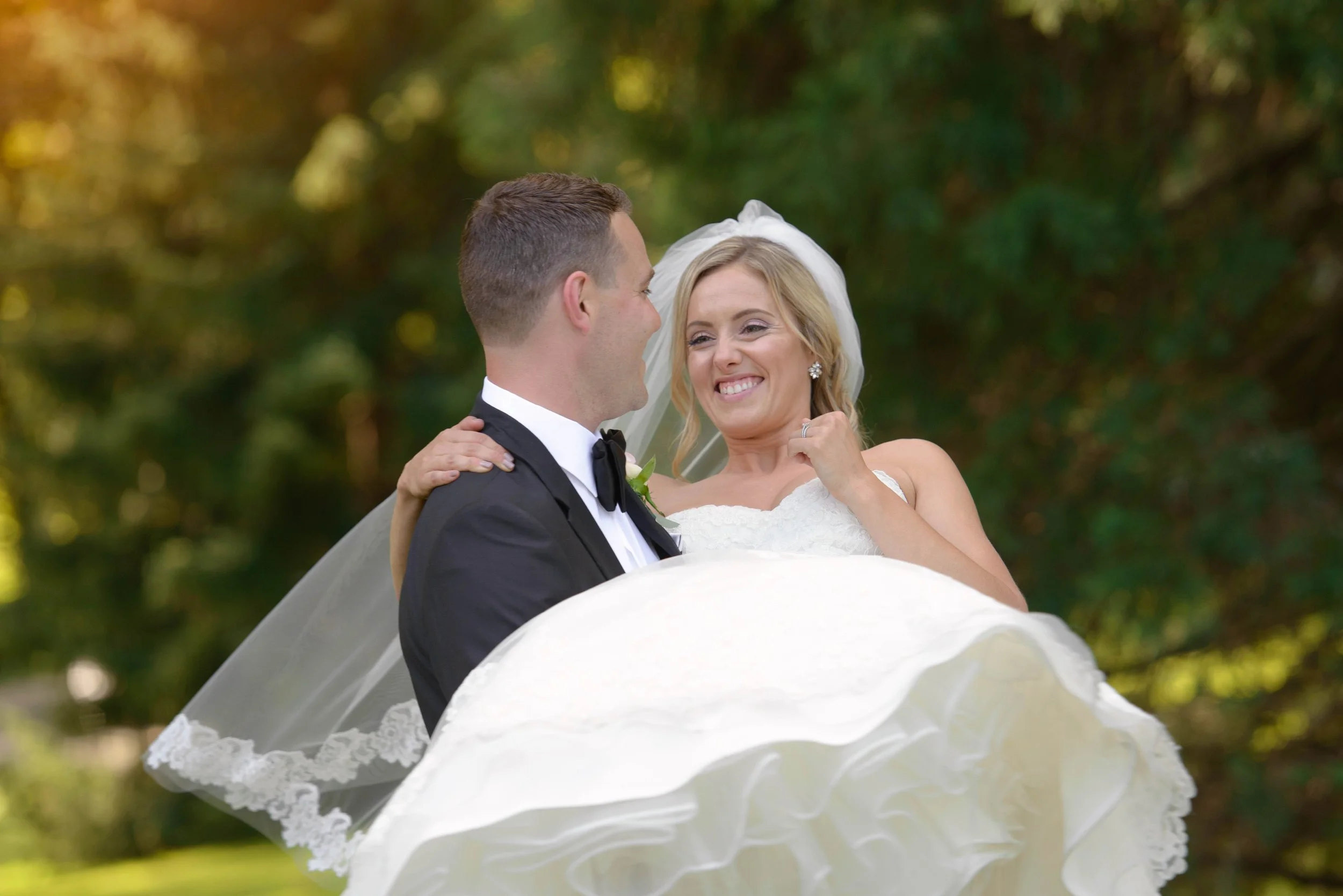 A bride and groom smiling as the groom lifts the bride outdoors, with green trees in the background.
