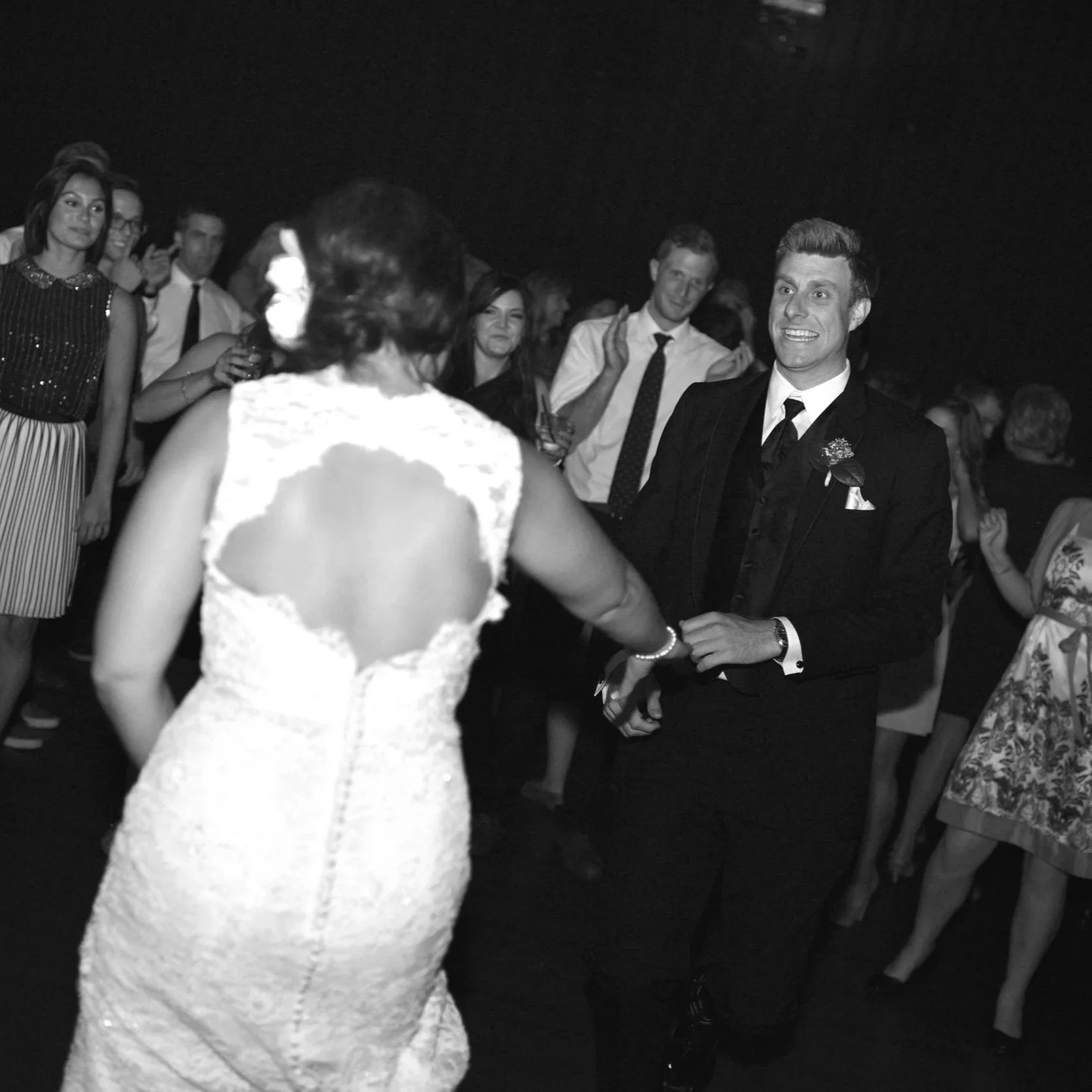 Black and white photo of a wedding reception with guests dancing and socializing. The bride is visible from behind in a lace dress, and the groom is smiling at her, dressed in a suit with a boutonniere.