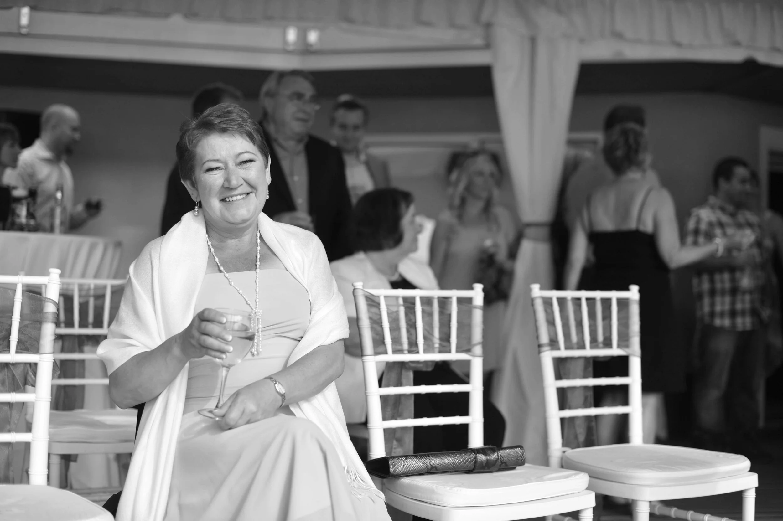 A smiling woman in formal attire sitting at a table, holding a glass of wine, at a social event with other guests in the background.