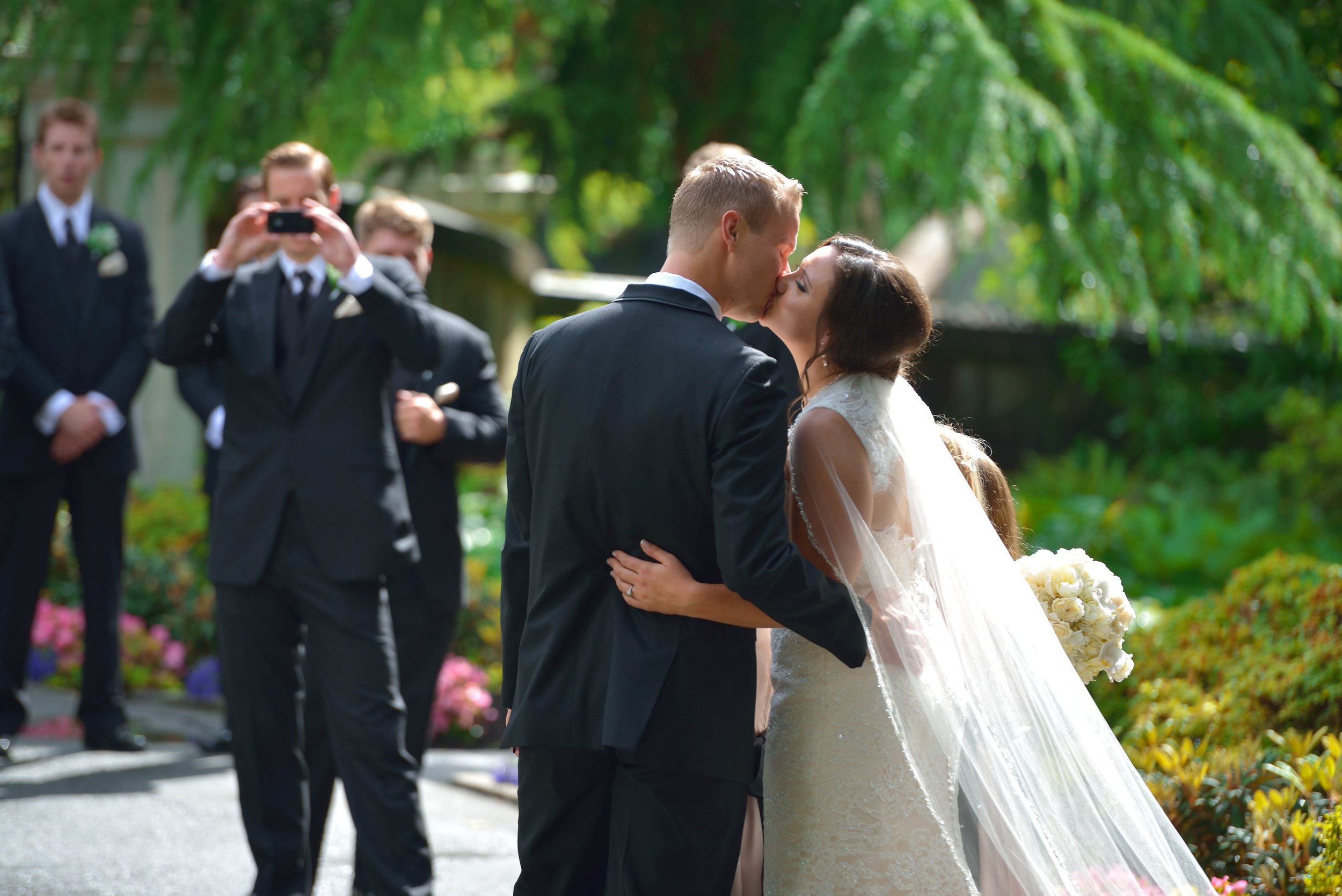 A bride and groom share a kiss at their wedding ceremony outdoors surrounded by greenery, while three groomsmen in tuxedos watch and take photos.