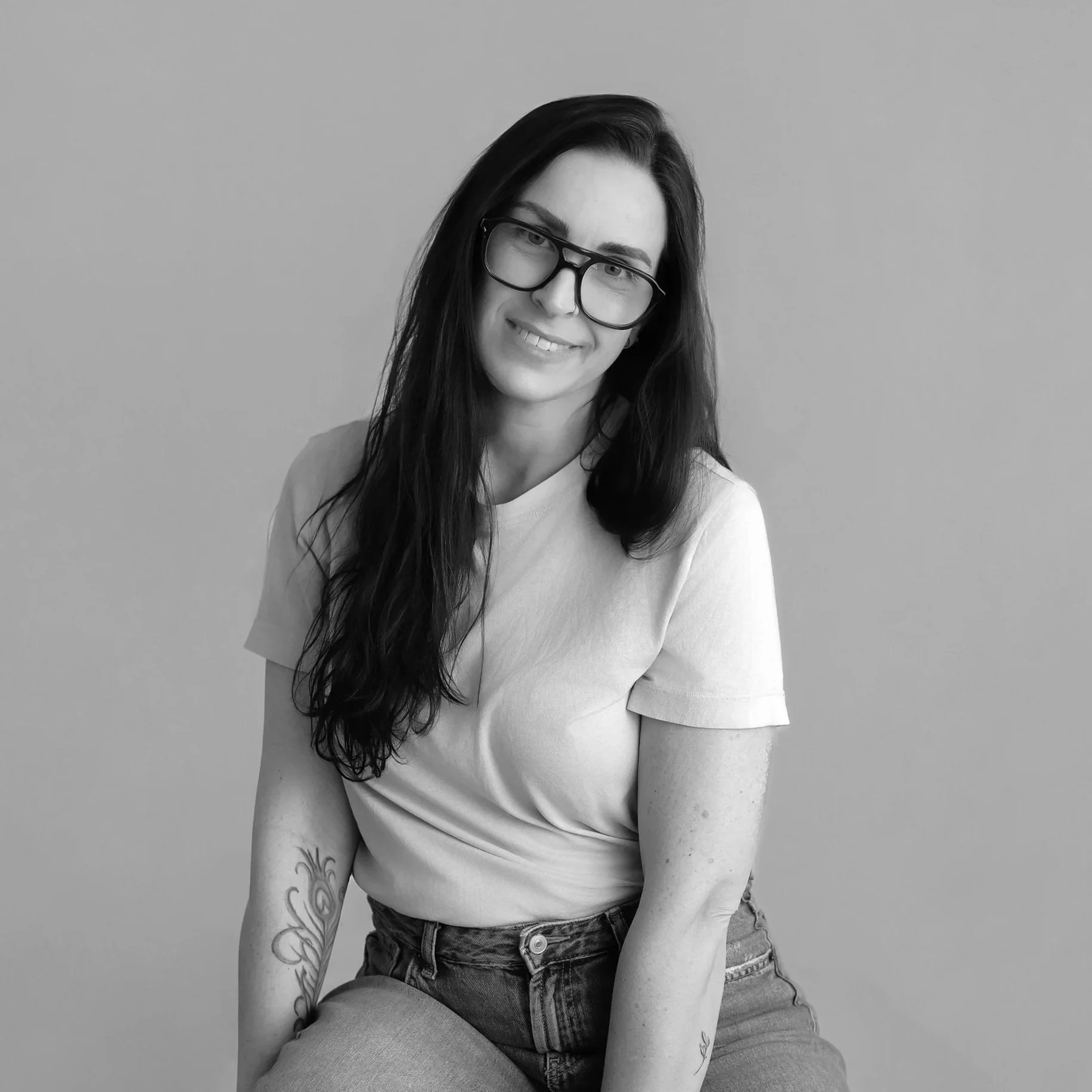 Black and white photo of a woman with long dark hair, wearing glasses and a light-colored t-shirt, sitting against a plain wall.