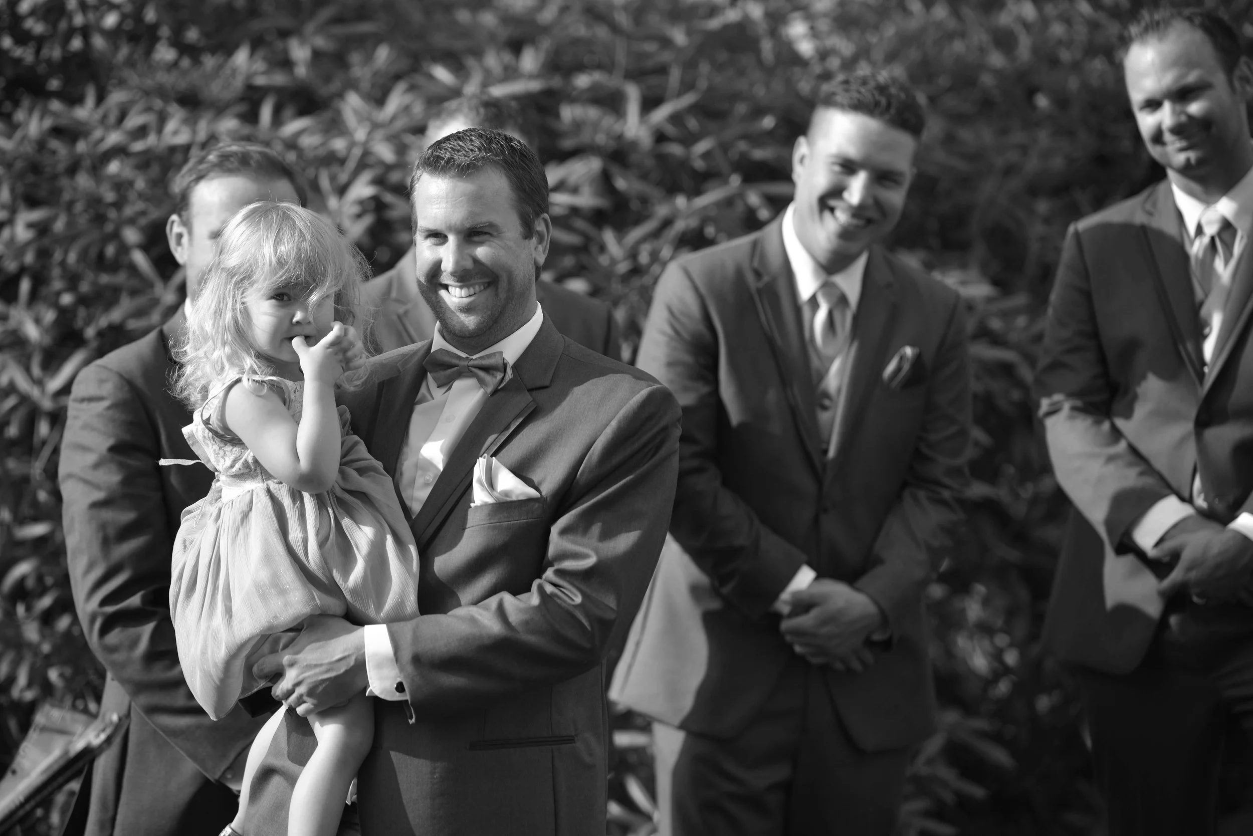 A group of young men in suits sitting outdoors, with a young girl in a dress with a bow, smiling and sitting on a man's lap.