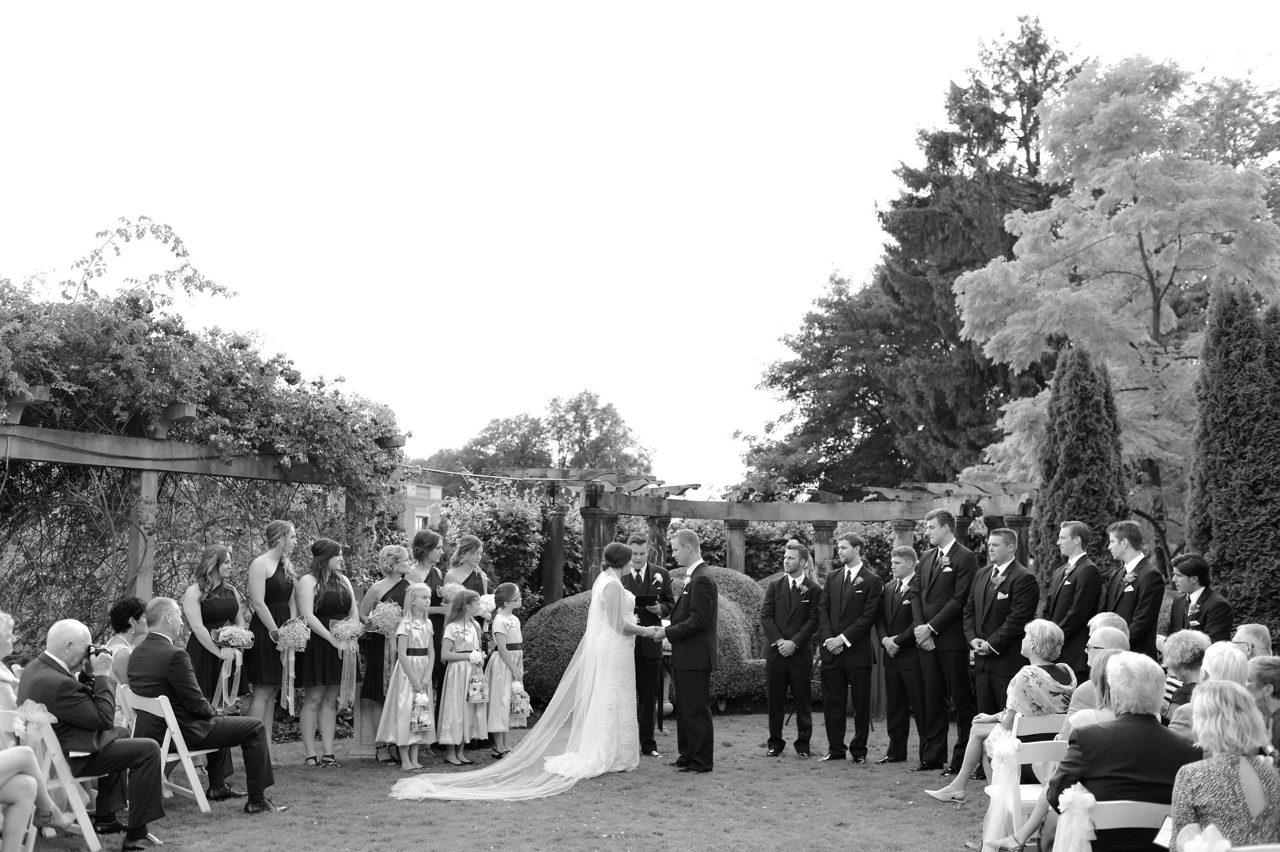 A wedding ceremony outdoors in a garden with trees and cloudy sky, bride and groom exchanging vows, surrounded by bridesmaids and groomsmen, guests seated on white chairs.