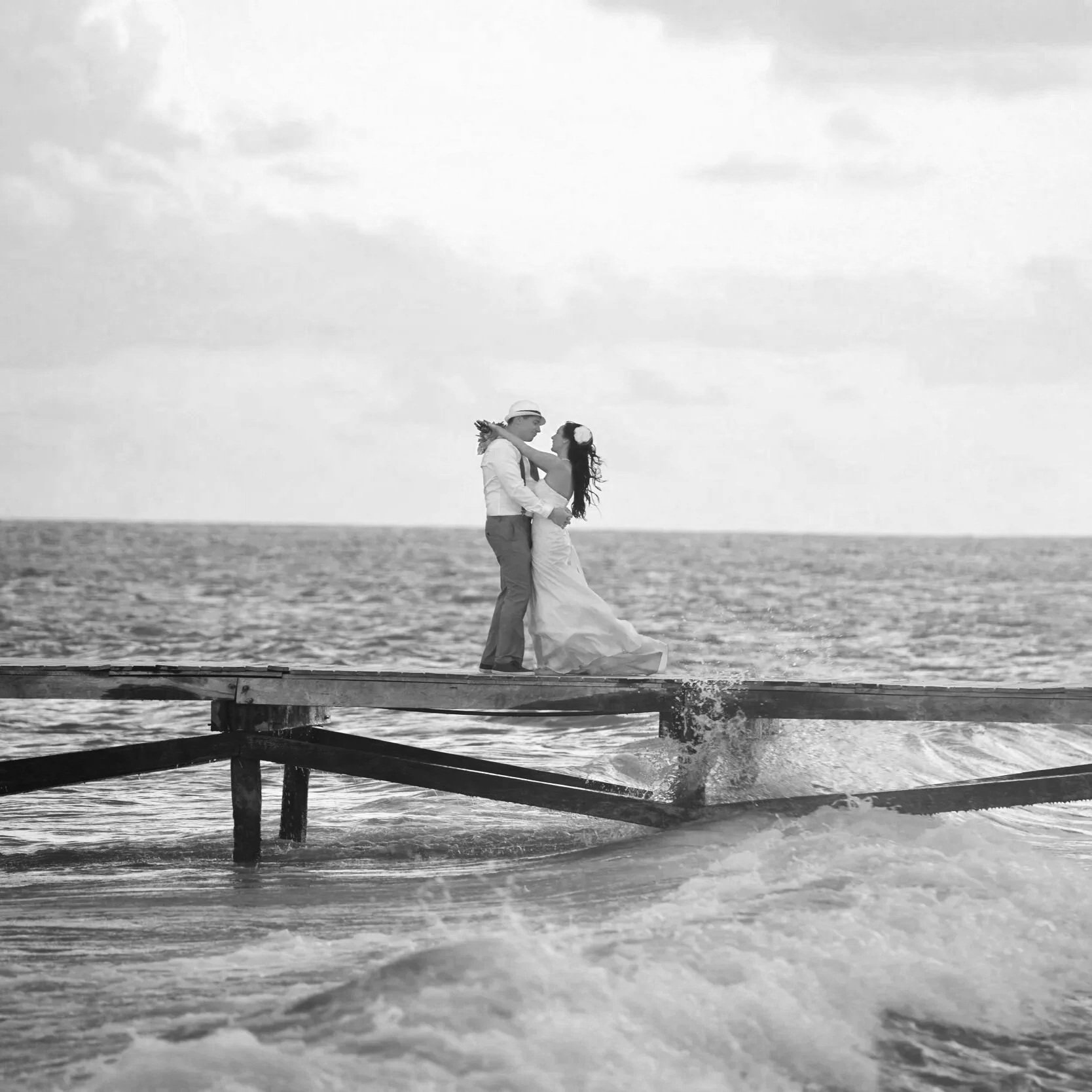 A black-and-white photo of a couple dressed in wedding attire, standing on a wooden dock at the beach, embracing each other with the ocean and cloudy sky in the background.