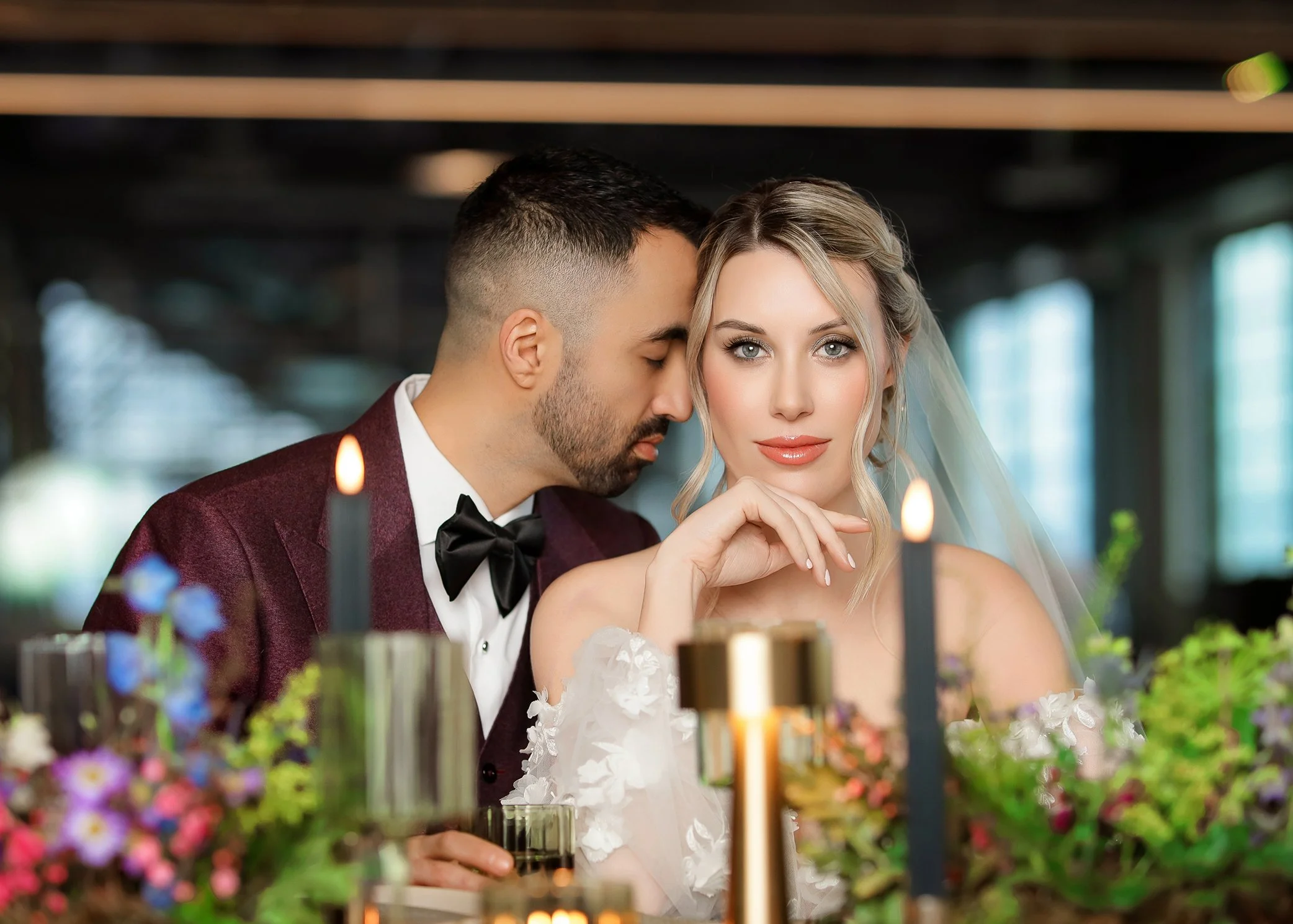 A couple dressed in wedding attire sitting at a table with candles and flowers, with the groom whispering into the bride's ear.