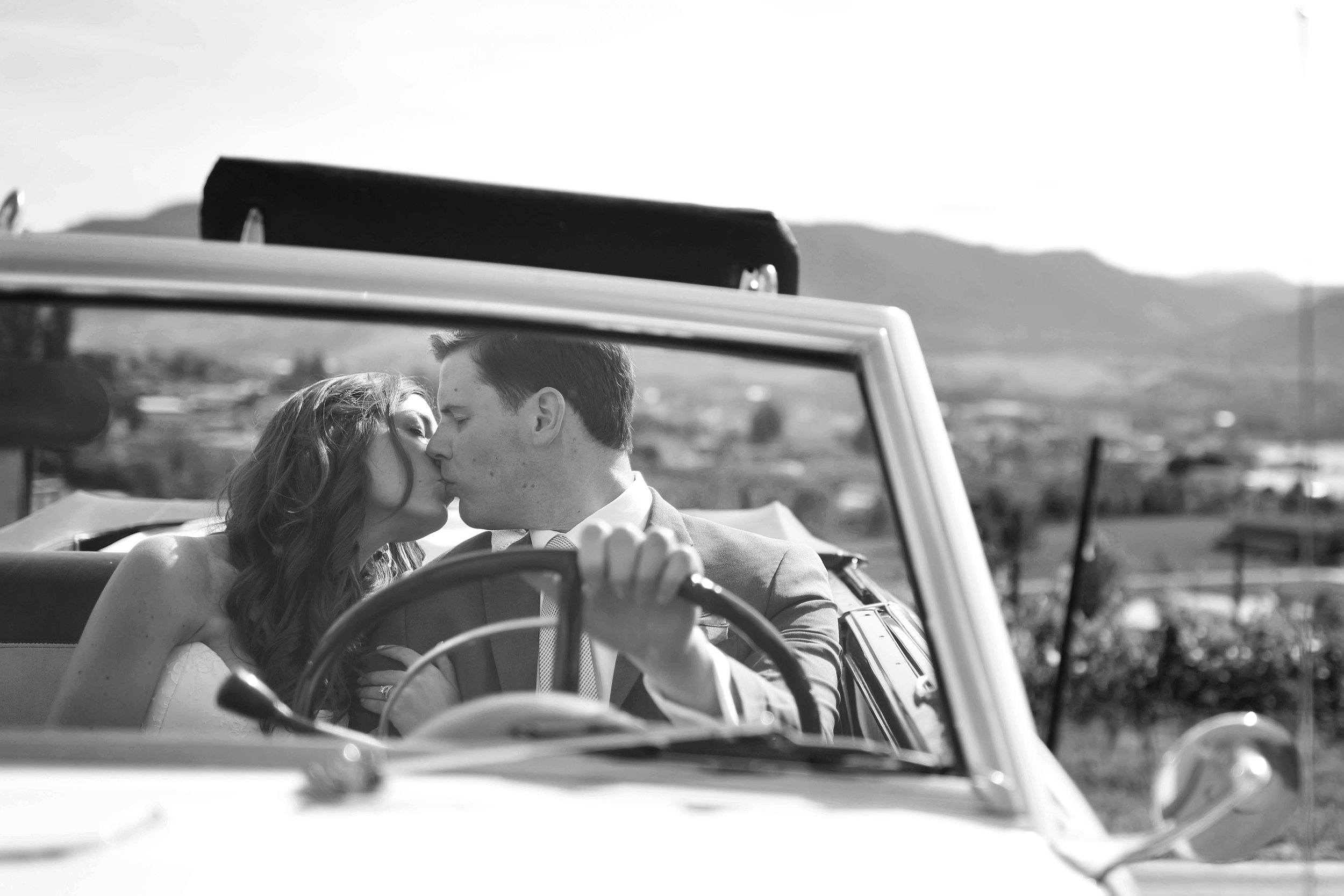 A black-and-white photo of a couple sharing a kiss while sitting in a vintage convertible car, with scenic landscape and mountains in the background.