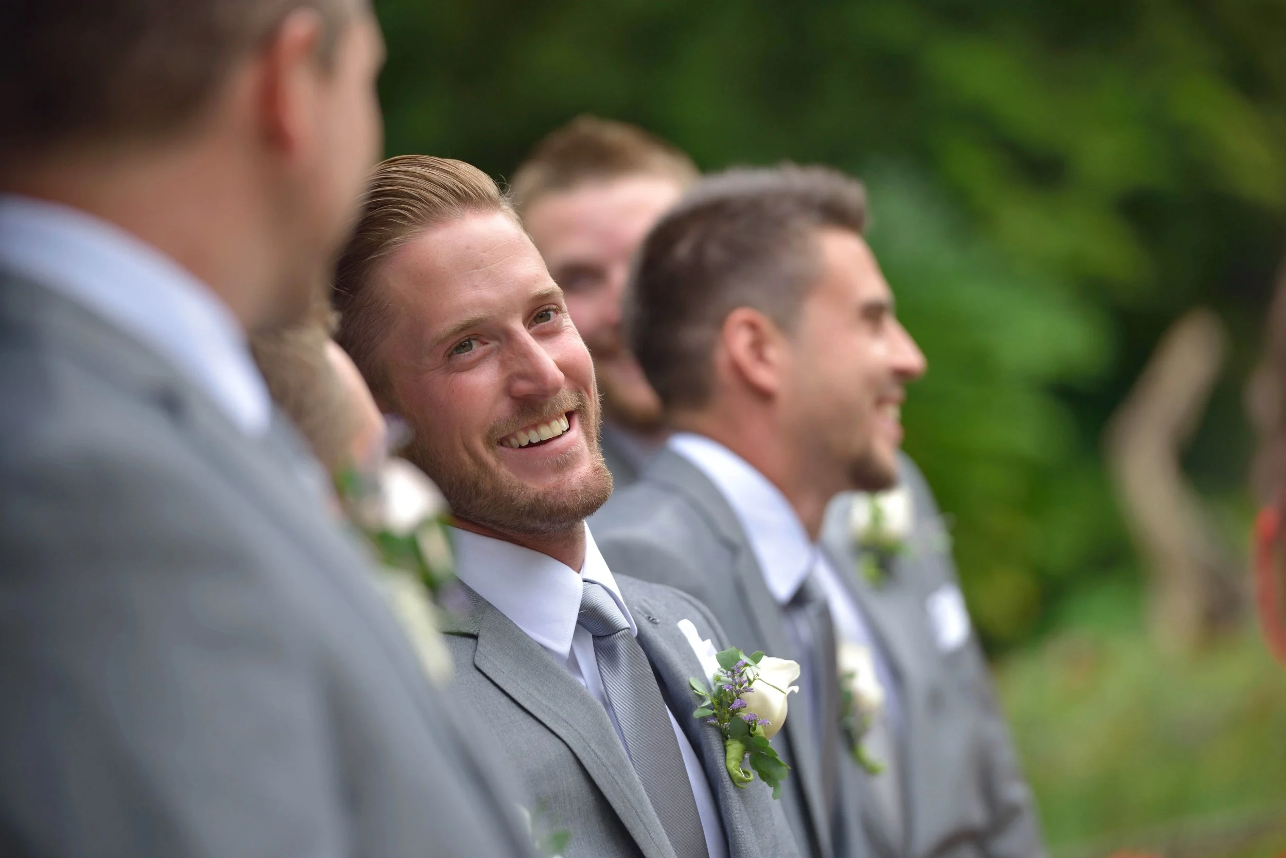 A group of men in gray suits and white shirts, standing in a row outdoors with greenery in the background. One man is smiling and looking to his right, with a boutonniere pinned to his suit.