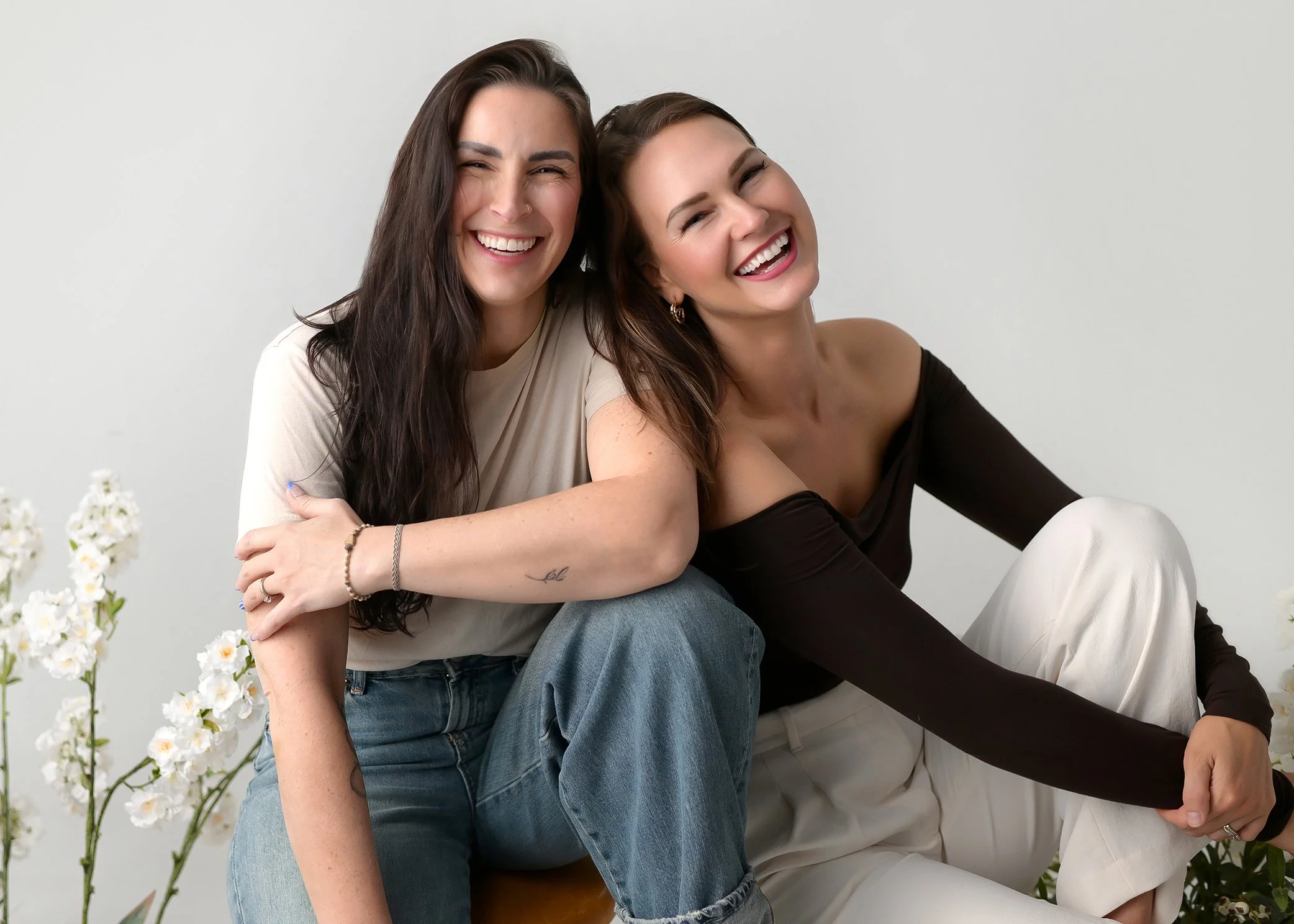 Two women smiling and sitting close together, with one woman sitting on the other's lap, against a plain background with white flowers on the side.
