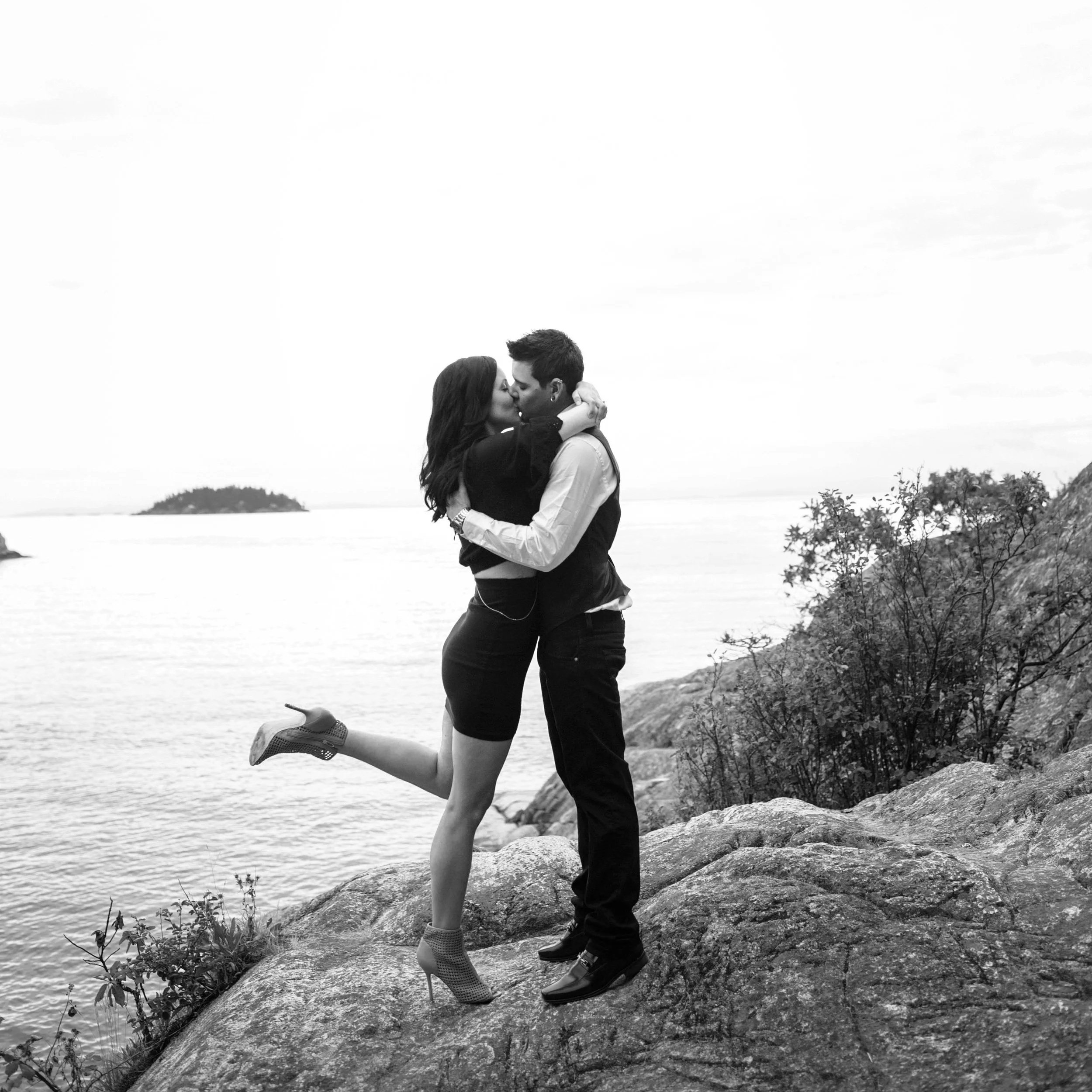 A couple kissing on a rock near the water, with the woman raising her leg and wearing heeled boots, the man in dress shoes, outdoors with trees and water in the background.