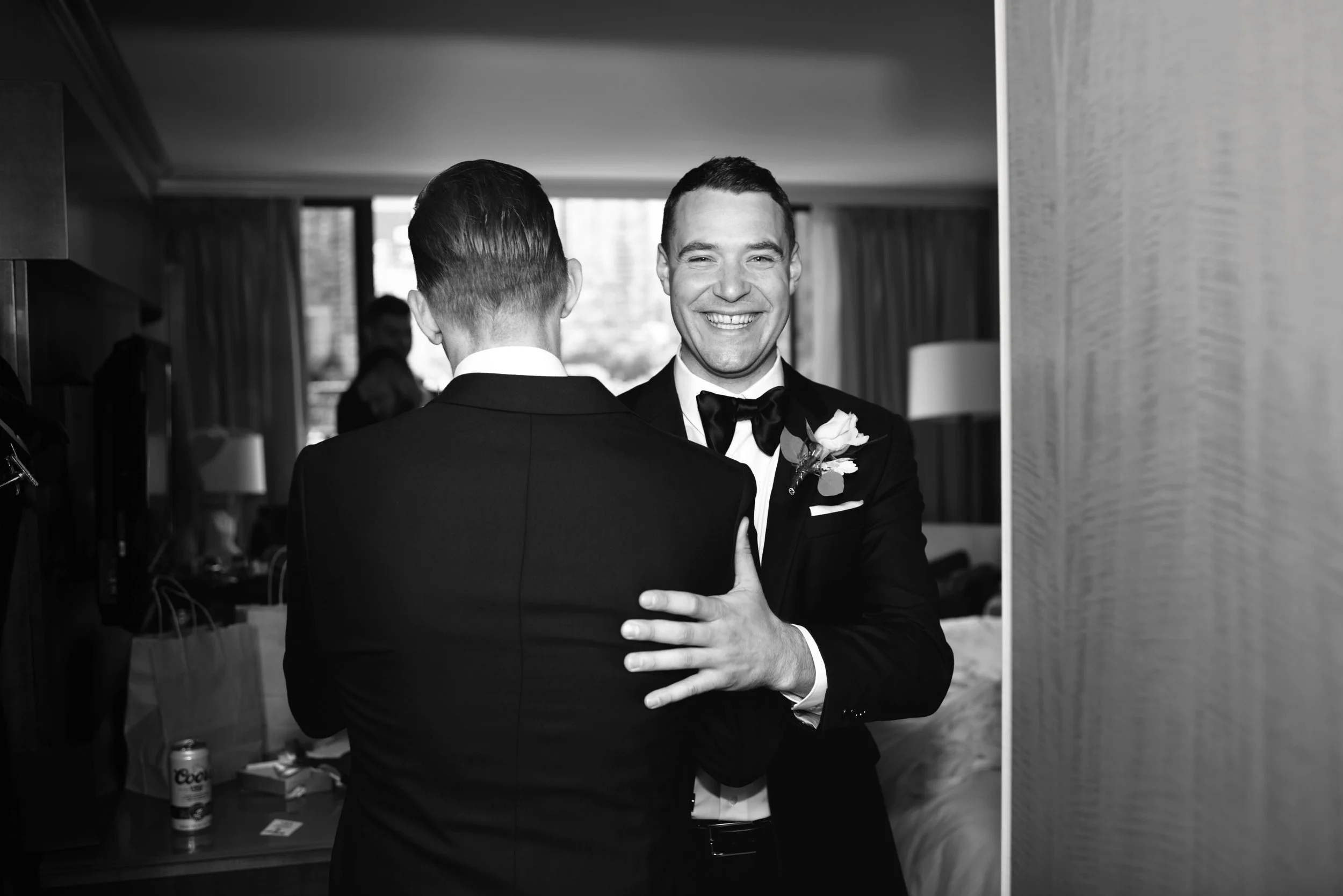 Two men in tuxedos embrace and smile at each other in an indoor setting, possibly at a formal event or wedding, with a window and curtains in the background.