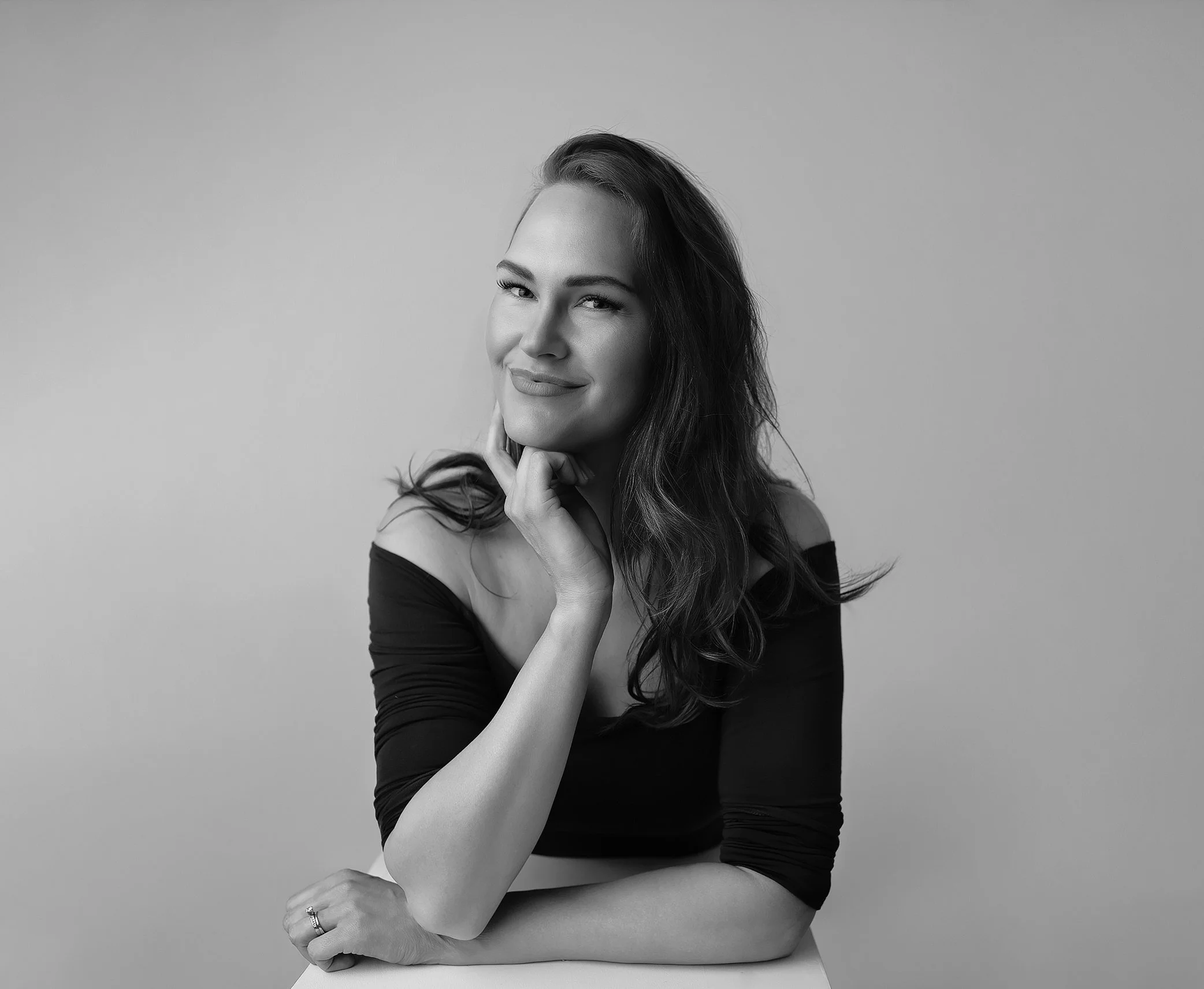 Black and white portrait of a woman with long hair, smiling slightly, with one hand touching her face, wearing an off-shoulder top.