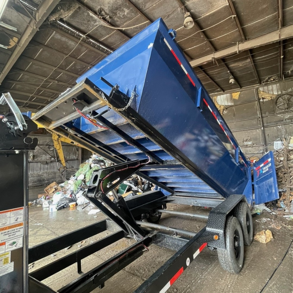 A large blue dump trailer attached to a tow vehicle, inside a warehouse or garage, with trash and debris on the ground.