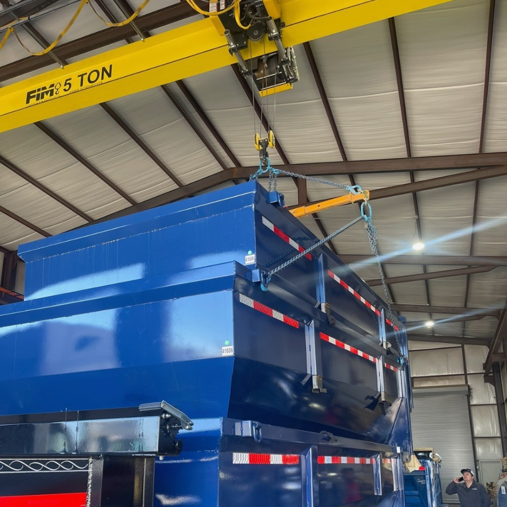 A large blue semi-truck trailer inside a warehouse being lifted by a yellow overhead crane.